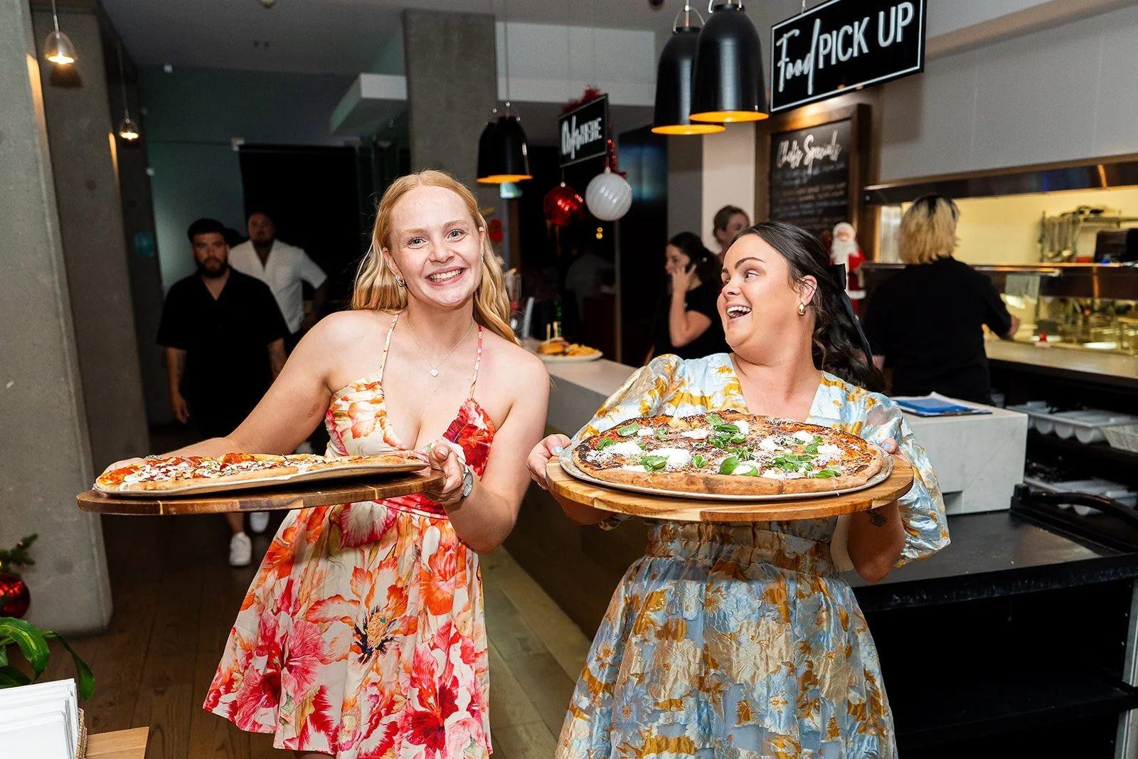 Two women wearing floral dresses, holding large pizza trays, smiling and enjoying their time in a pizzeria. The background shows a restaurant interior with a sign that says 'To Go/Takeout Pickup' and several other people.