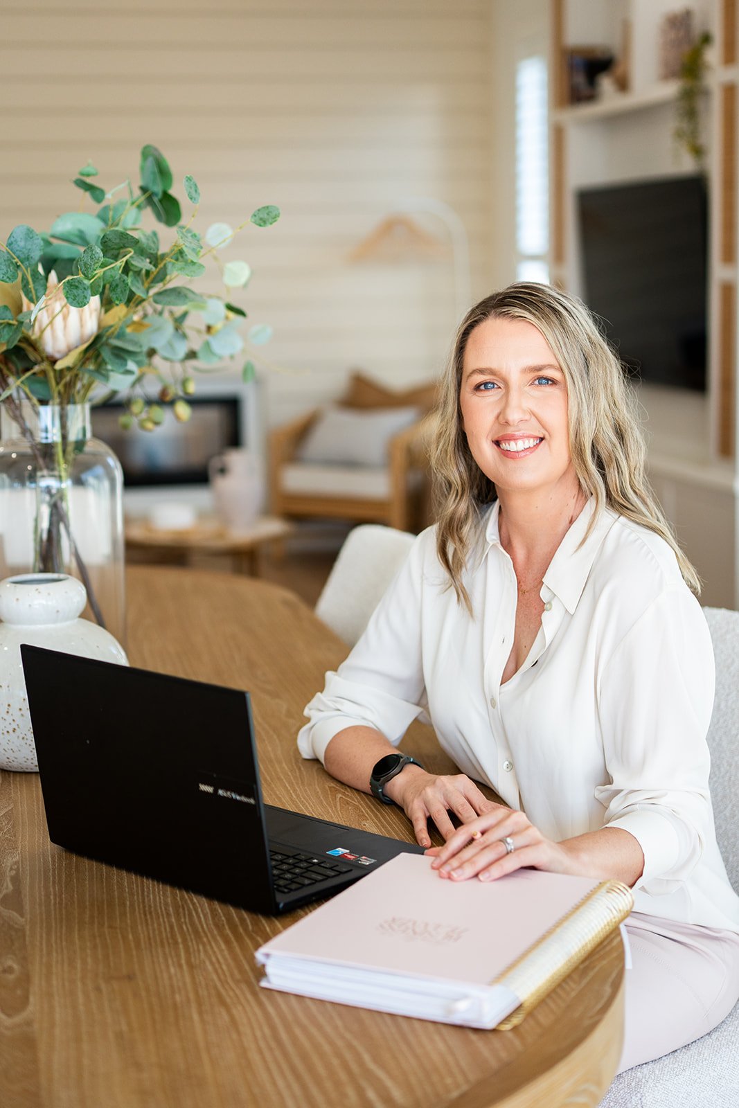 A smiling woman in a white shirt sitting at a wooden table with a laptop, notebook, and a large vase with greenery and a flower inside, in a cozy, well-lit living room.