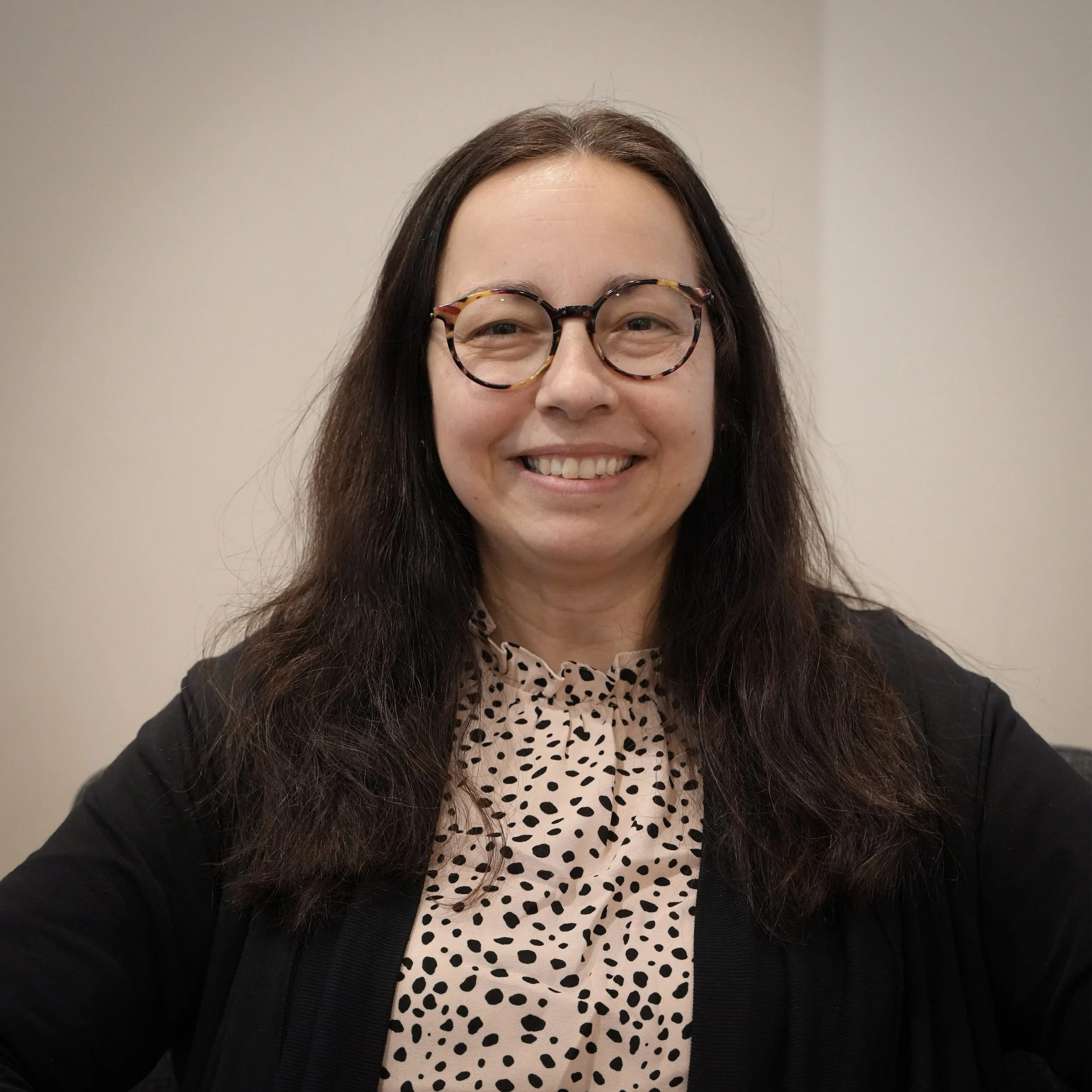 A woman with dark brown hair, wearing glasses and a black blazer, smiling in front of a plain beige wall.