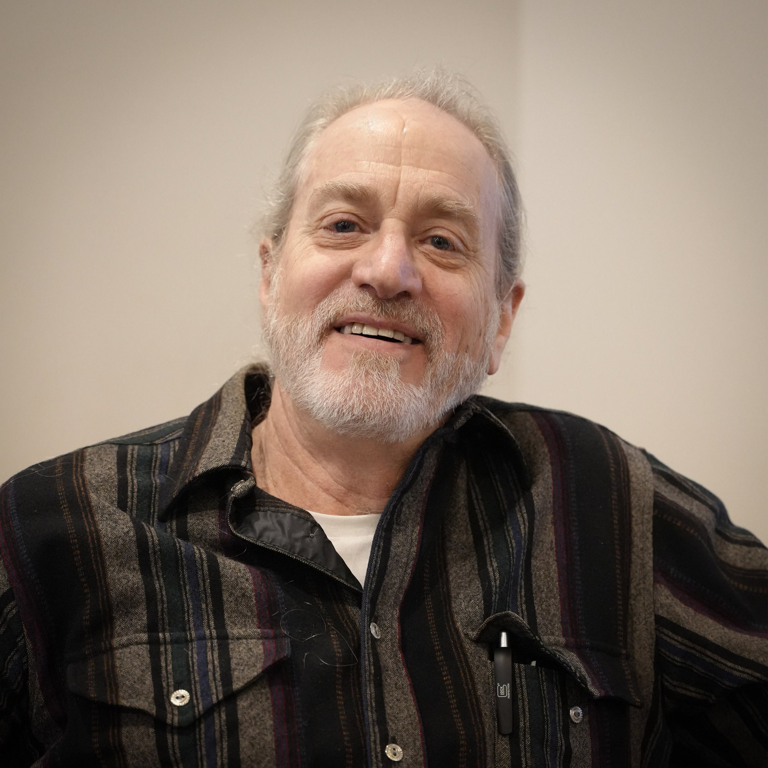 Portrait of an elderly man with gray hair and beard, smiling, wearing a striped shirt, against a plain light-colored background.