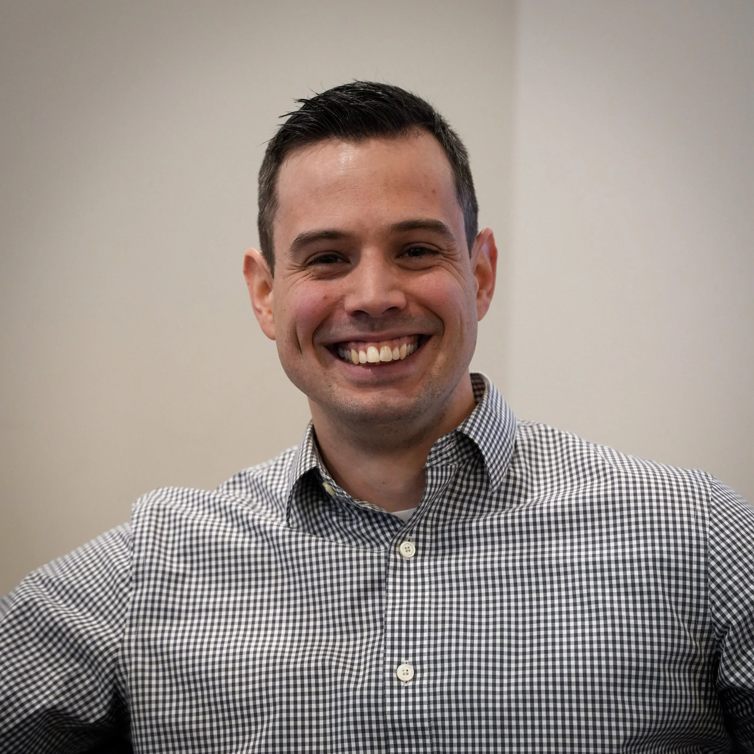 A smiling man with short dark hair wearing a black and white checkered shirt against a plain background.