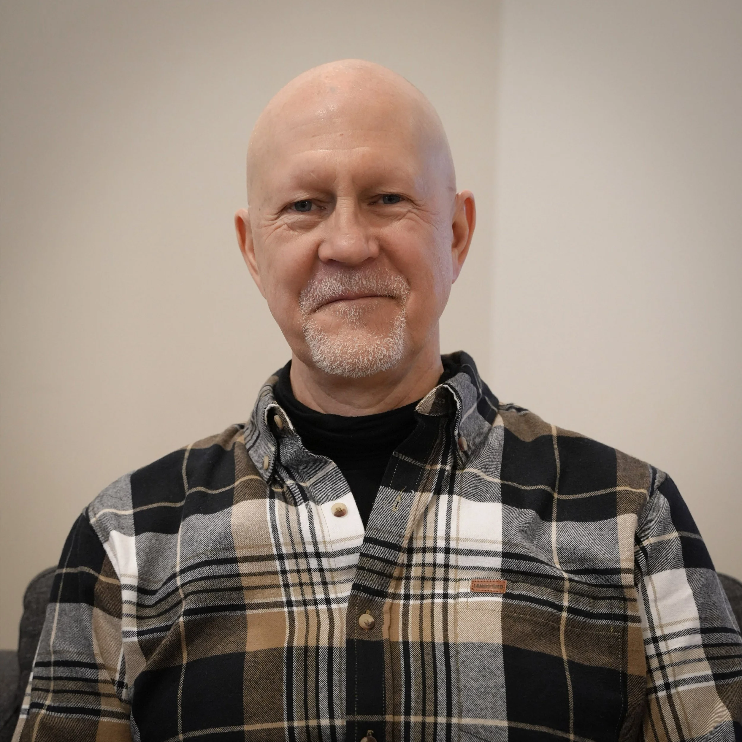 A middle-aged man with a bald head and a goatee, wearing a black, white, and tan plaid shirt over a black turtleneck, standing indoors against a plain light-colored wall.