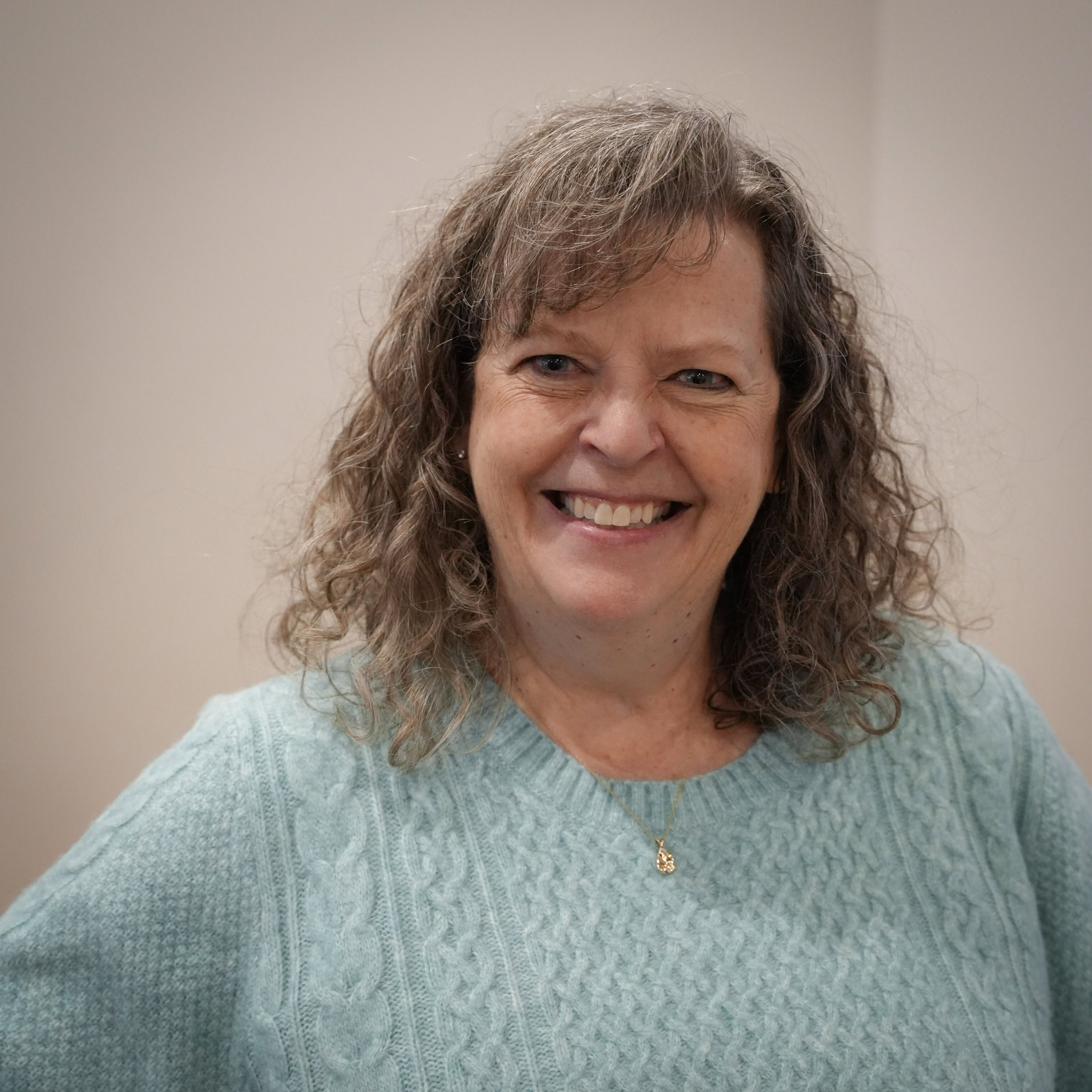A middle-aged woman with curly brown hair smiling, wearing a light blue sweater and a gold necklace, standing against a plain beige wall.
