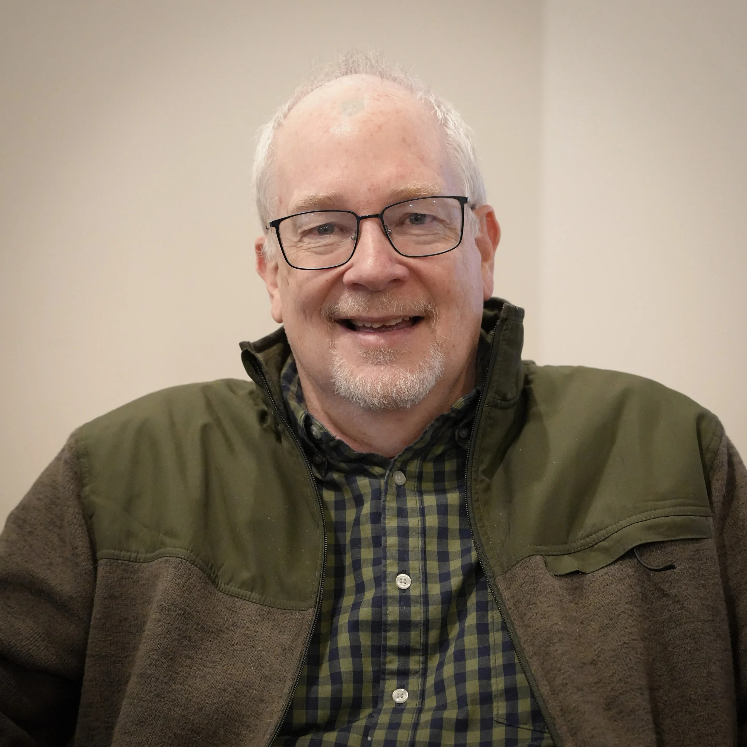 A smiling middle-aged man with glasses, white hair, and a beard, wearing a green jacket over a plaid shirt, sitting indoors against a plain wall.