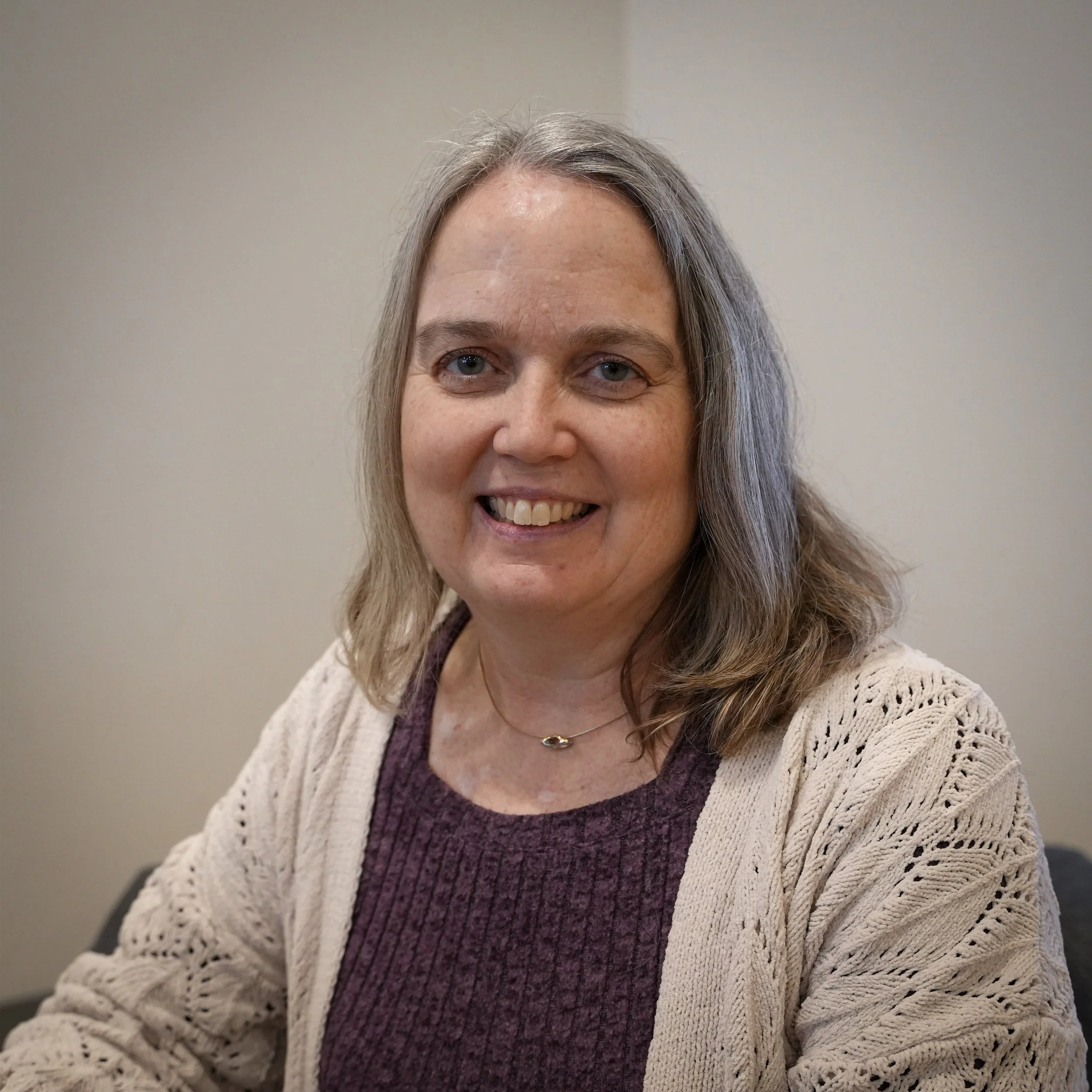 A smiling woman with shoulder-length gray hair wearing a beige knitted cardigan over a purple top.