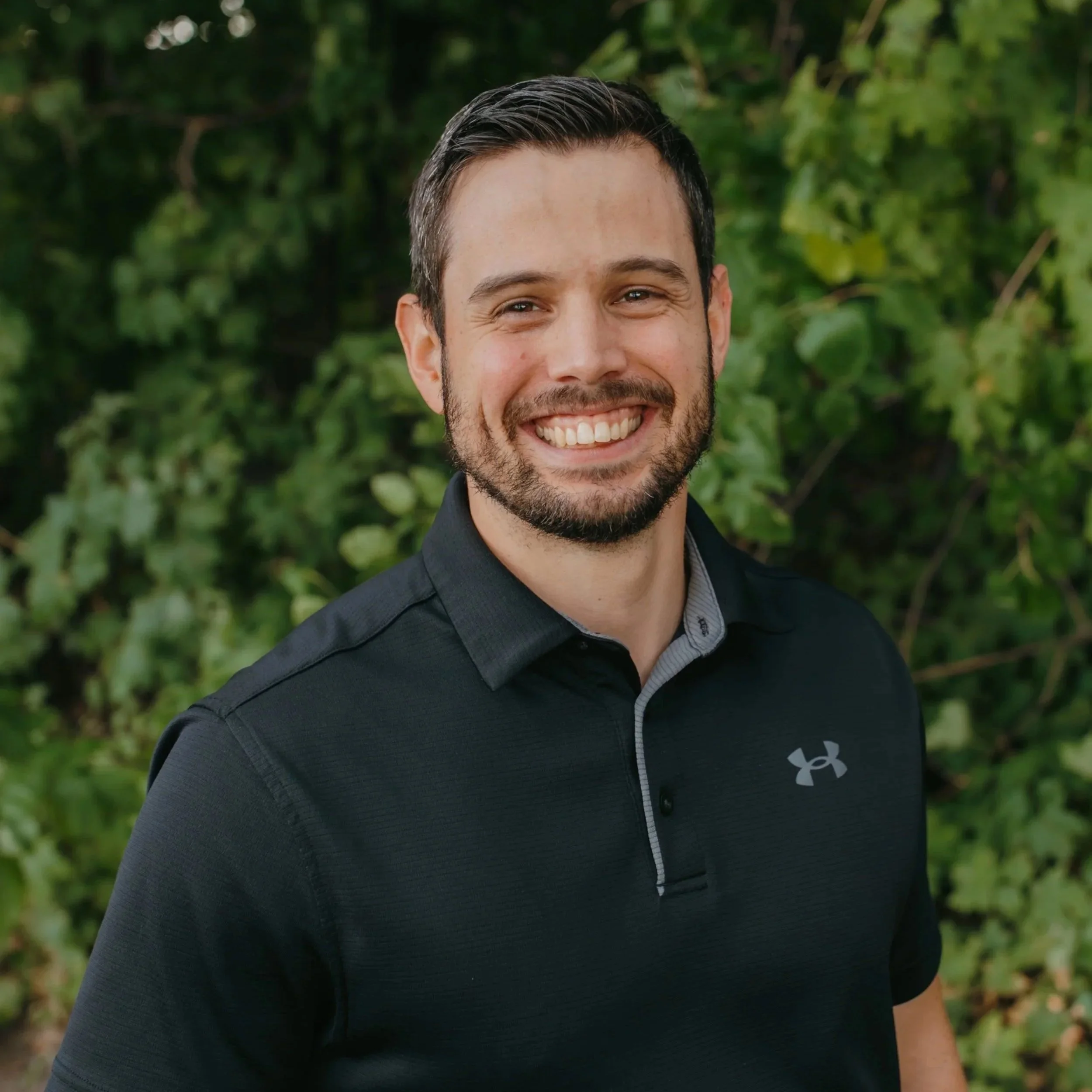 A smiling man with dark hair and a beard, wearing a black Under Armour polo shirt, standing outdoors in front of green foliage.