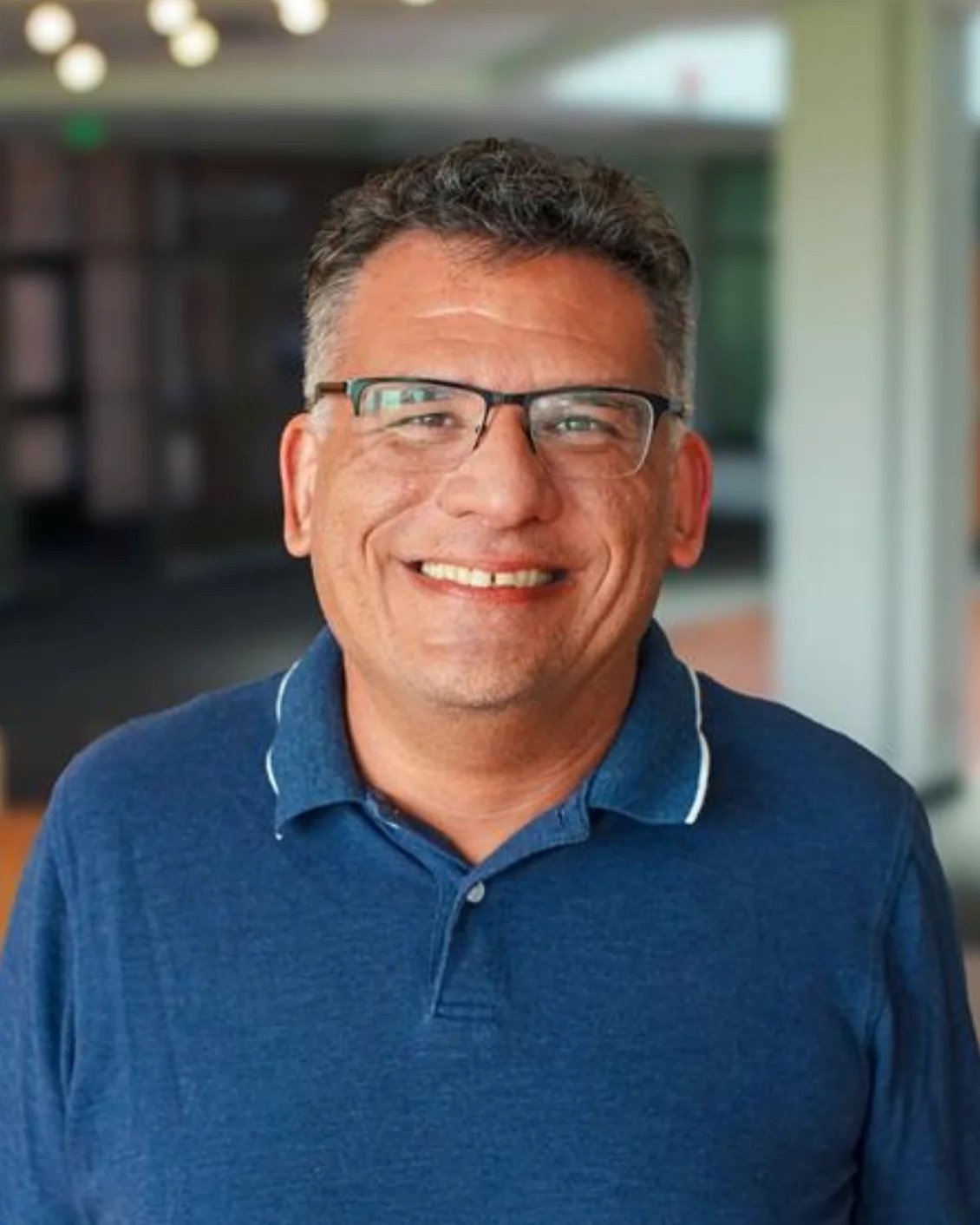 A middle-aged man with glasses and short curly hair, smiling, wearing a blue collared shirt, in an indoor setting with blurred background.