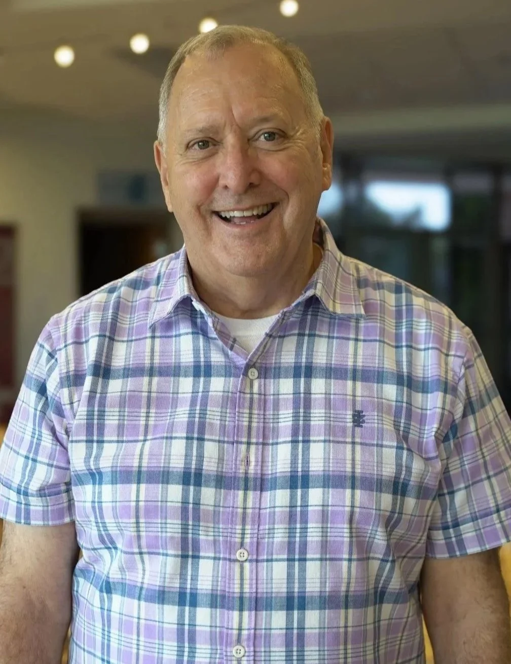 A smiling middle-aged man with short gray hair, wearing a colorful plaid shirt, standing indoors with blurred background.