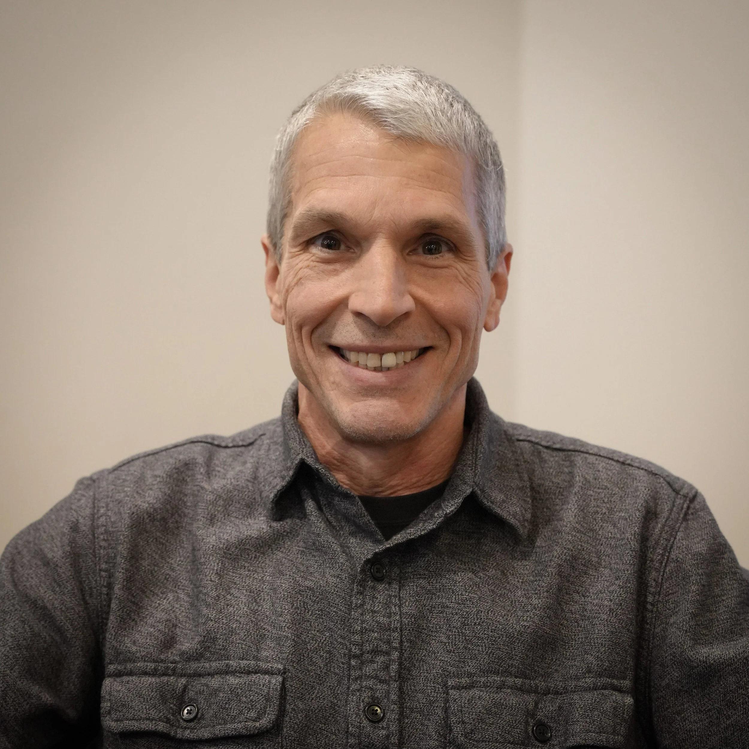 A smiling middle-aged man with gray hair wearing a dark gray button-up shirt against a plain light-colored background.