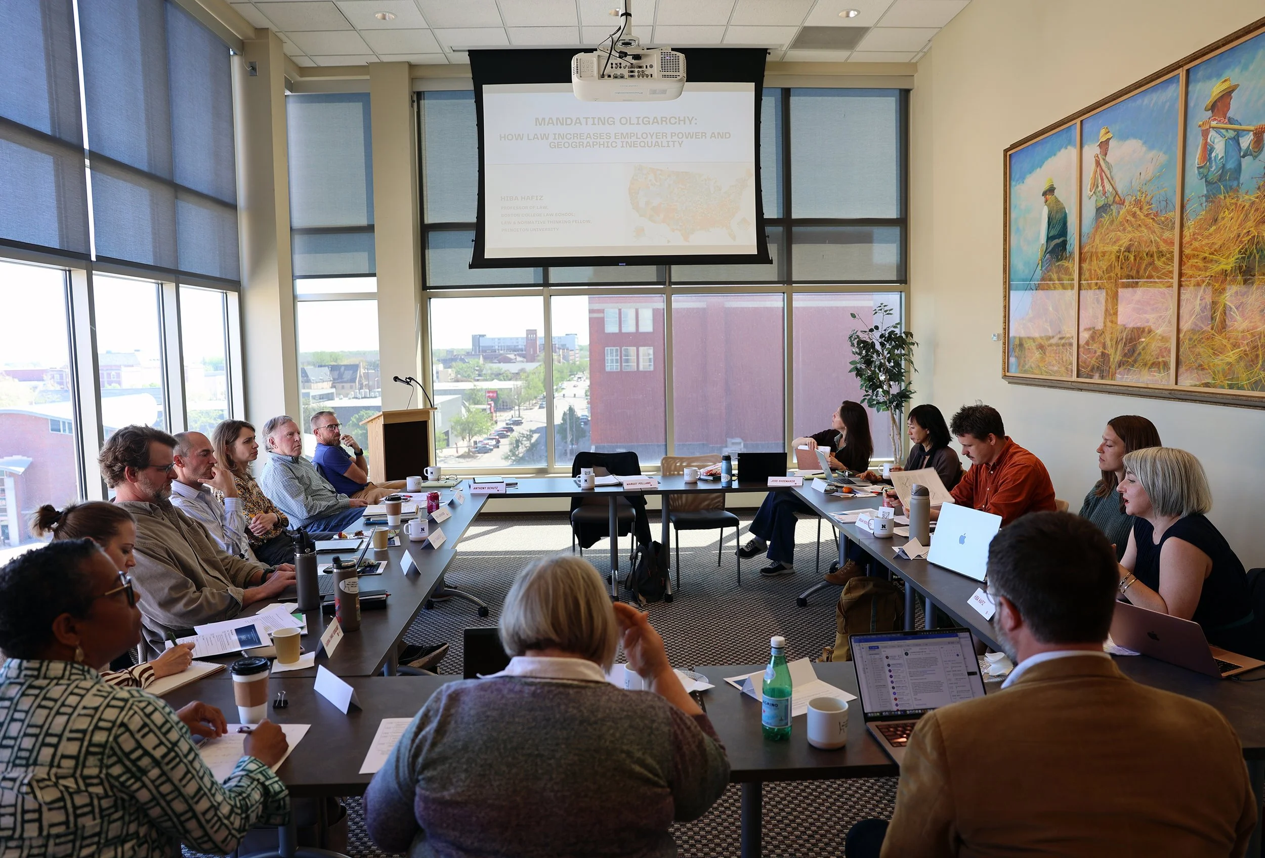 Photograph of a window-lined workshop room, with participants sitting around a table in shared discussion. Overhead, a slide authored by Hiba Hafiz is projected on the screen, showing a map of the United States and announcing the title of Hafiz's