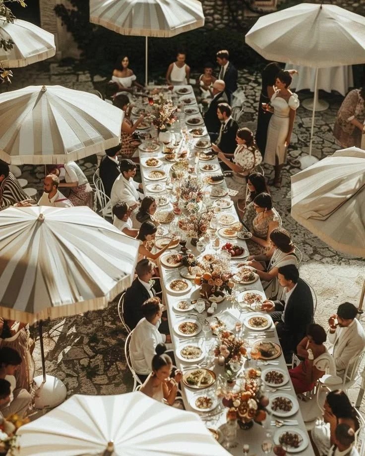An outdoor wedding reception with a long dining table set with plates and flowers, surrounded by guests and large white umbrellas.