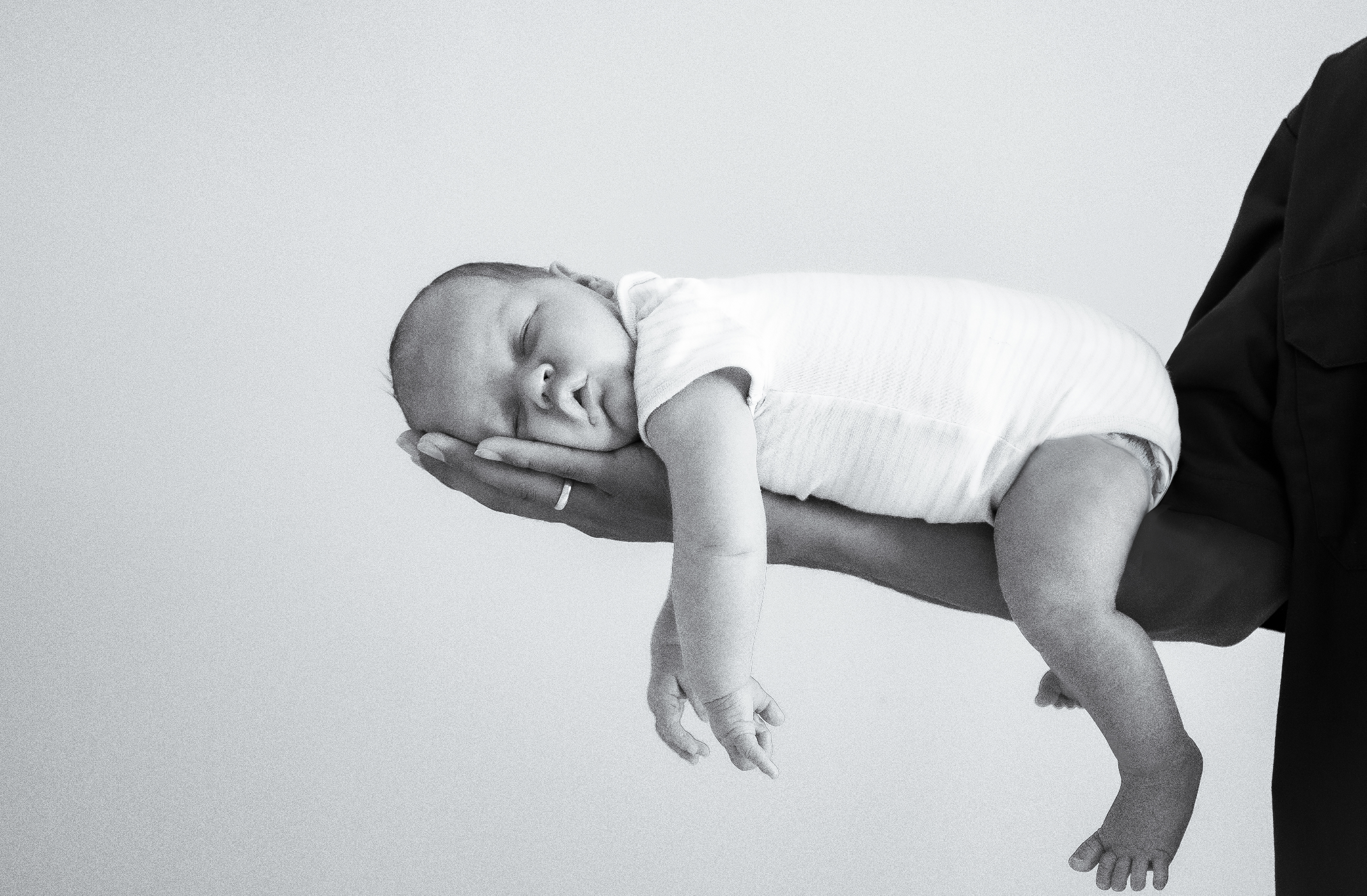 A newborn baby sleeping peacefully on an adult's hand, lying horizontally with arms and legs relaxed, in a black and white photo.