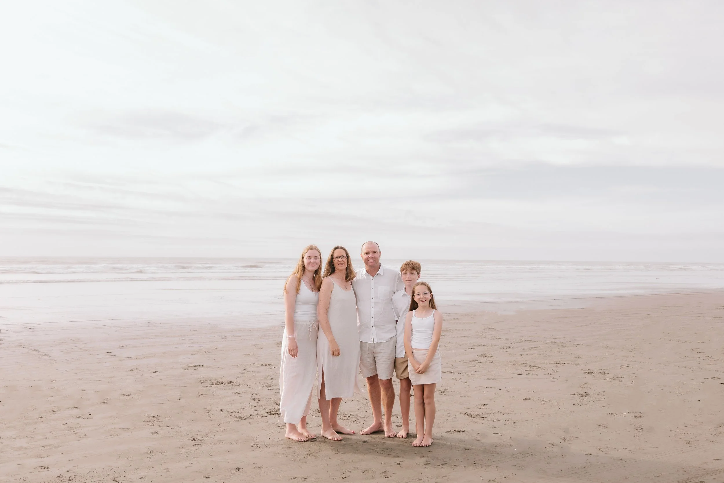 Family of five standing barefoot on the beach, dressed in white, with ocean and cloudy sky in the background.