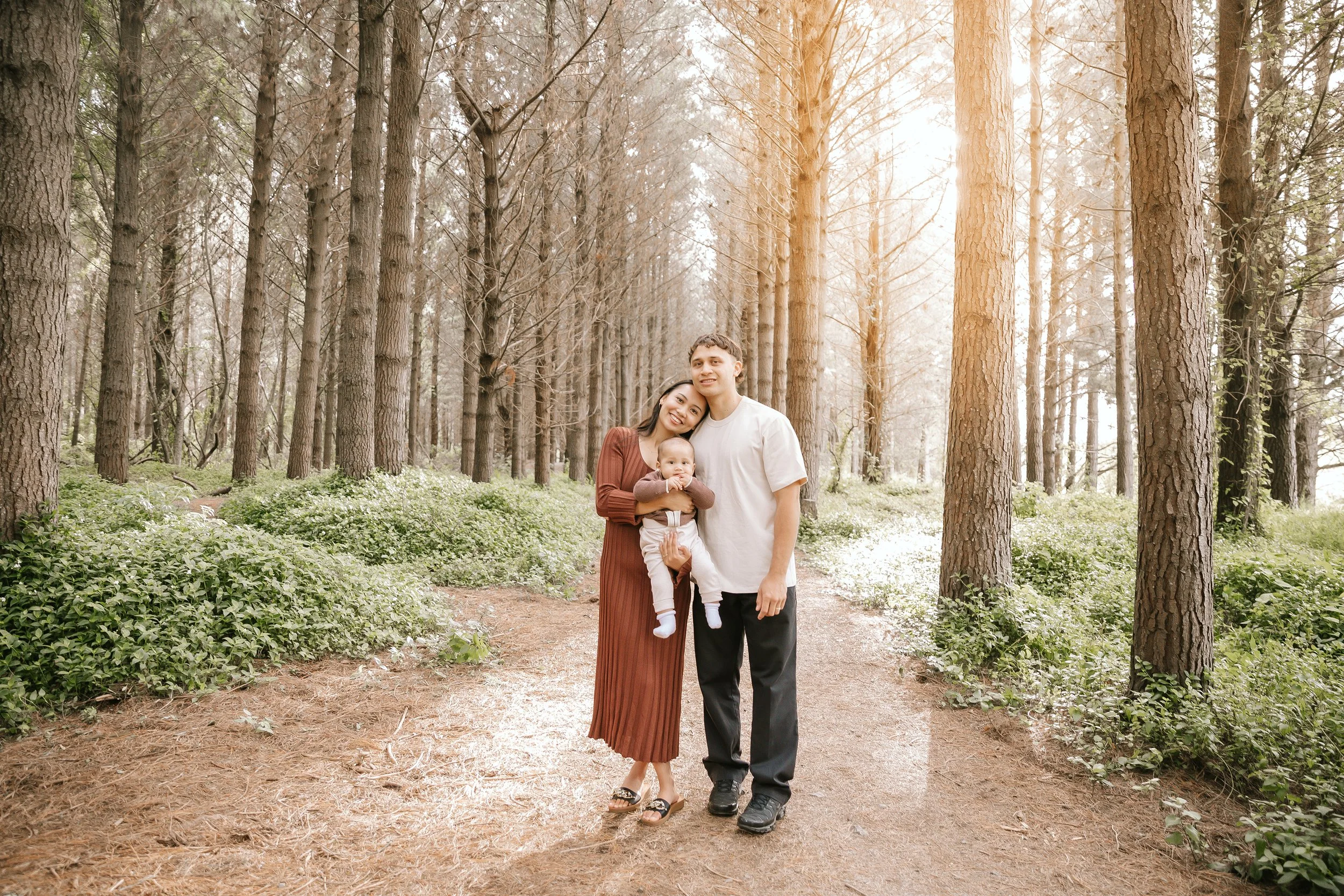 A family of three standing on a wooded forest path with tall trees and greenery, sunlight shining through the trees, the mother holding a baby, and the father standing beside them.