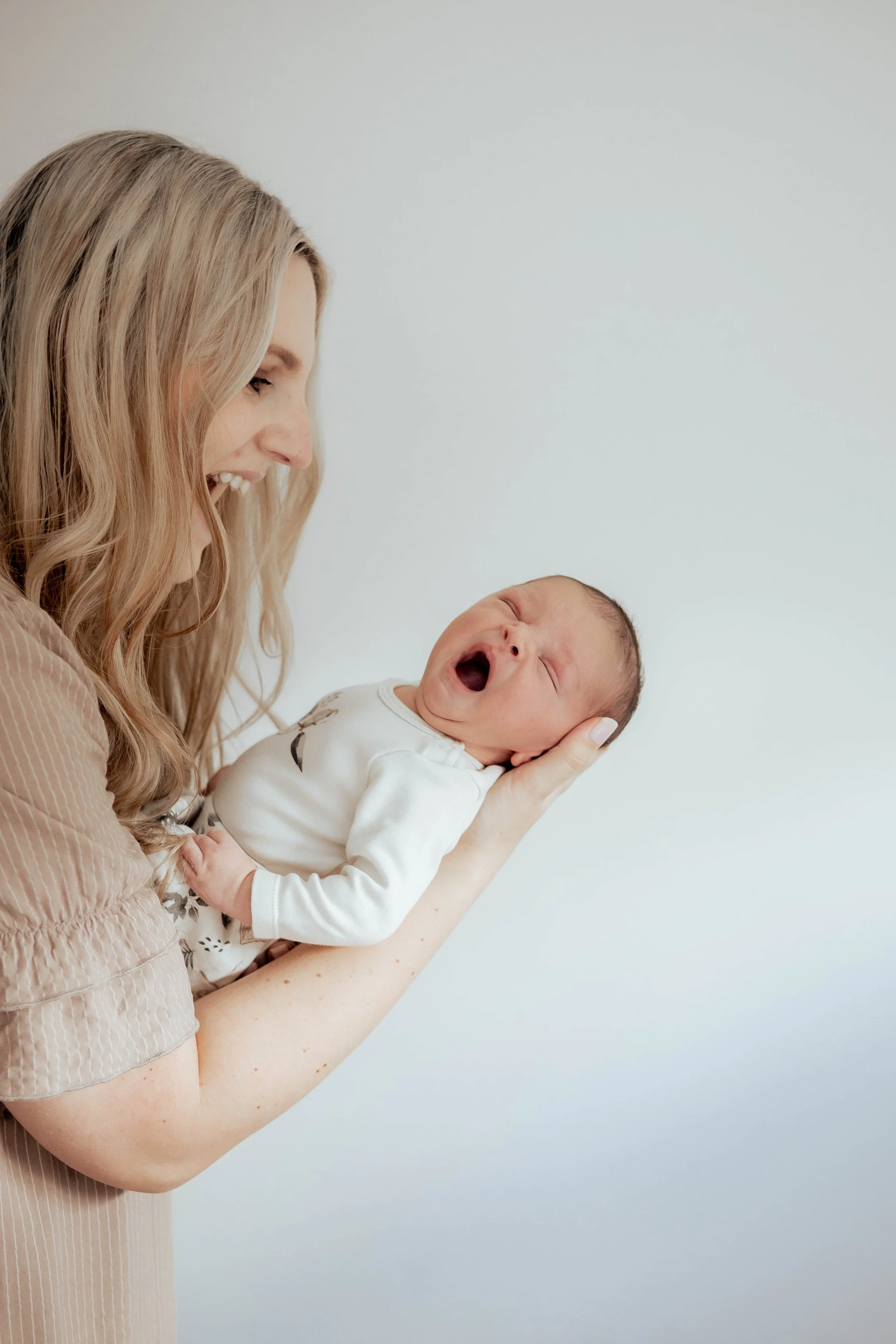 A woman holding a yawning baby near her face against a plain light background.