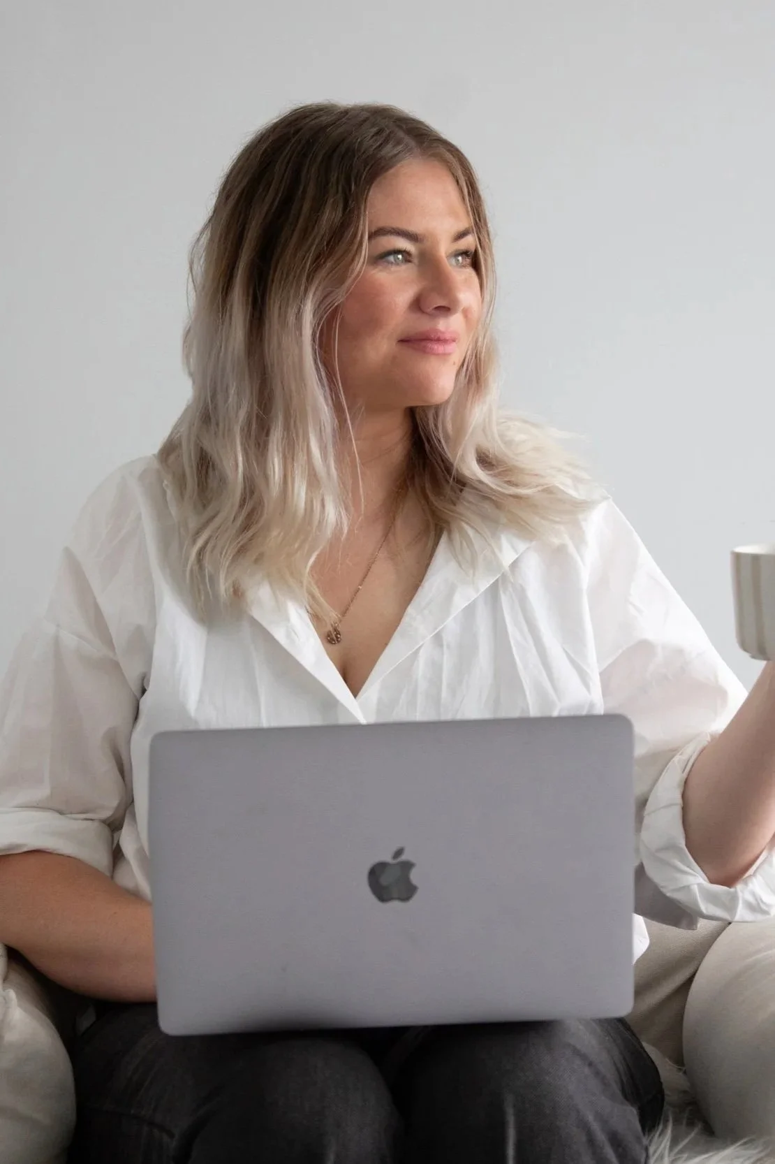 A woman with blonde wavy hair sitting on a couch, using a silver MacBook, holding a mug in her right hand, wearing a white shirt.