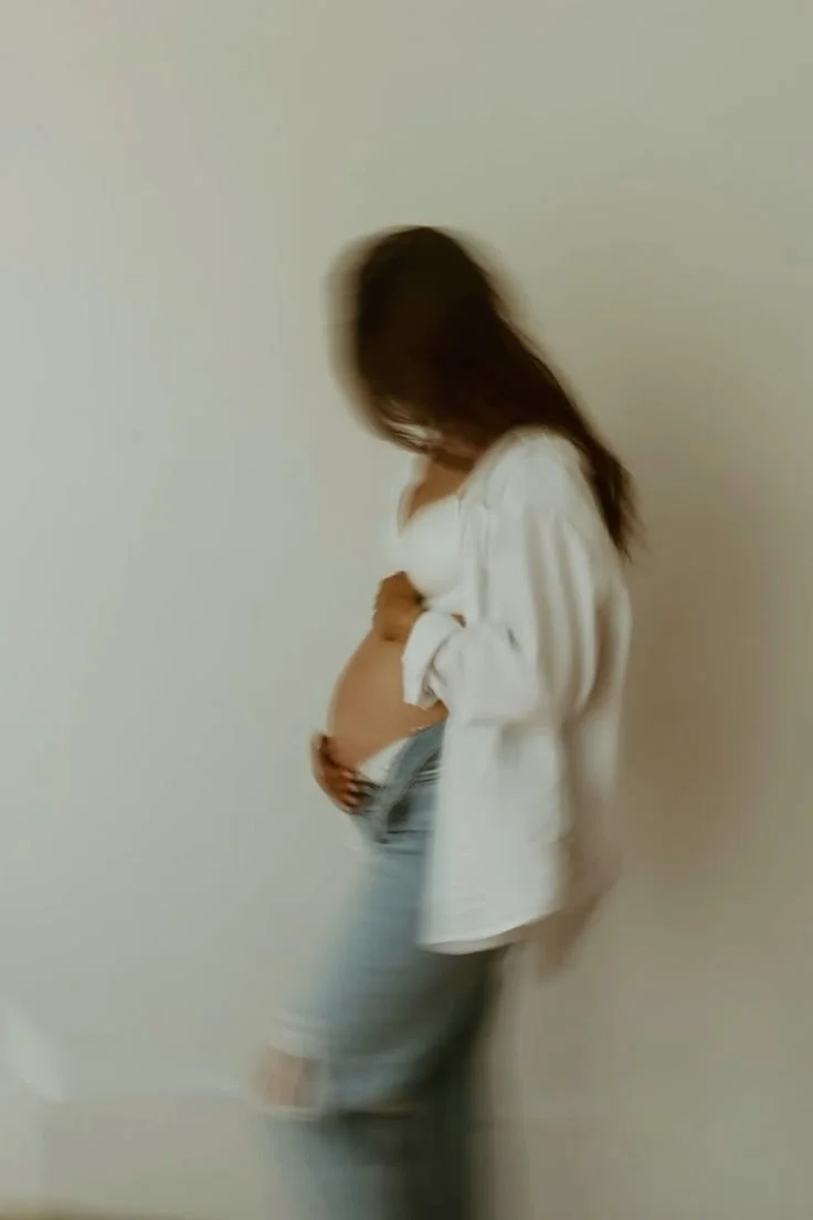 A woman standing against a plain wall, looking down and holding her pregnant belly with one hand.