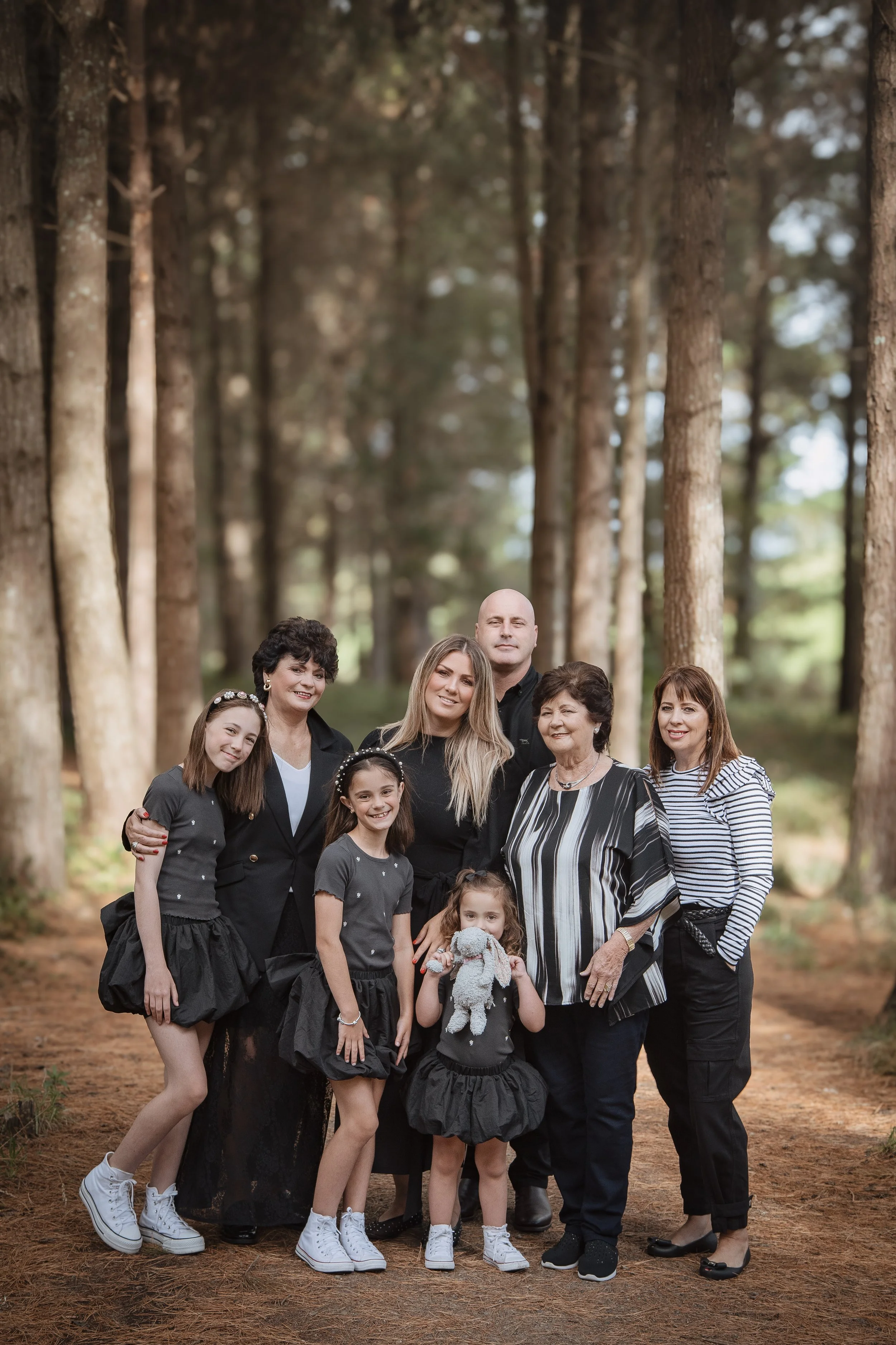 A multigenerational family group portrait outdoors in a forest with tall trees, including three men, four women, and three young girls, all dressed in coordinated dark and striped clothing.