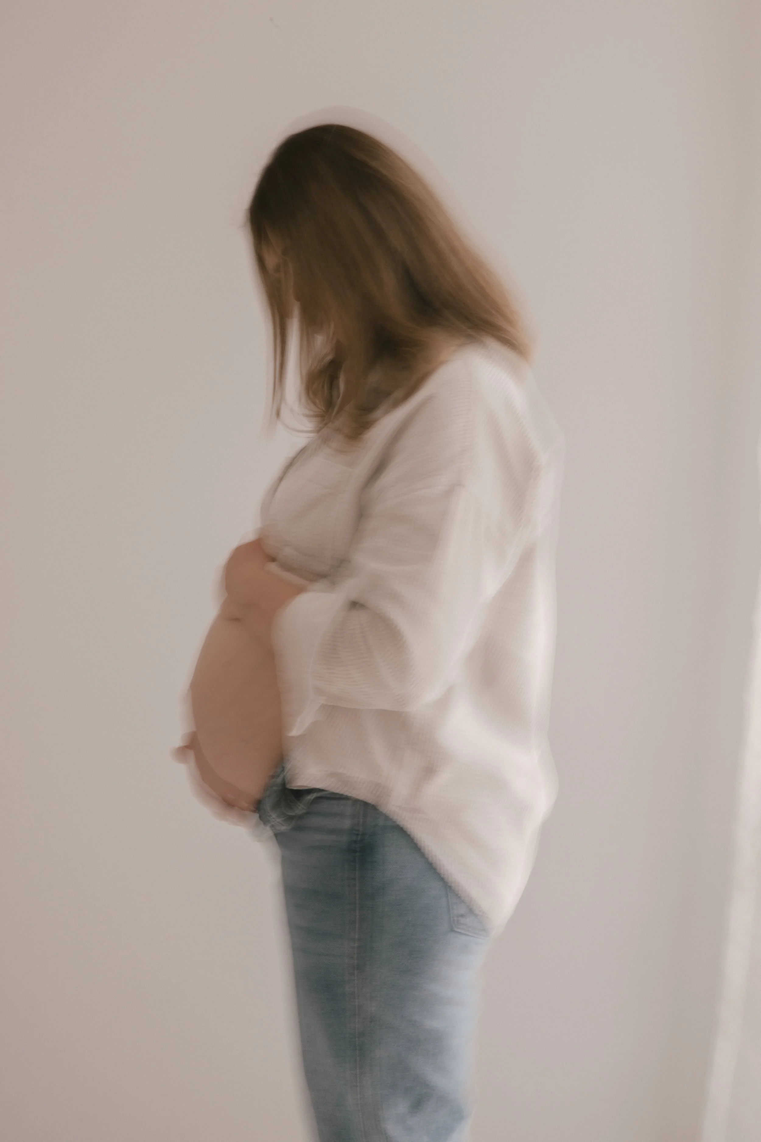 A woman with brown hair standing sideways, wearing a white long-sleeve top and blue jeans, gently holding her pregnant belly.