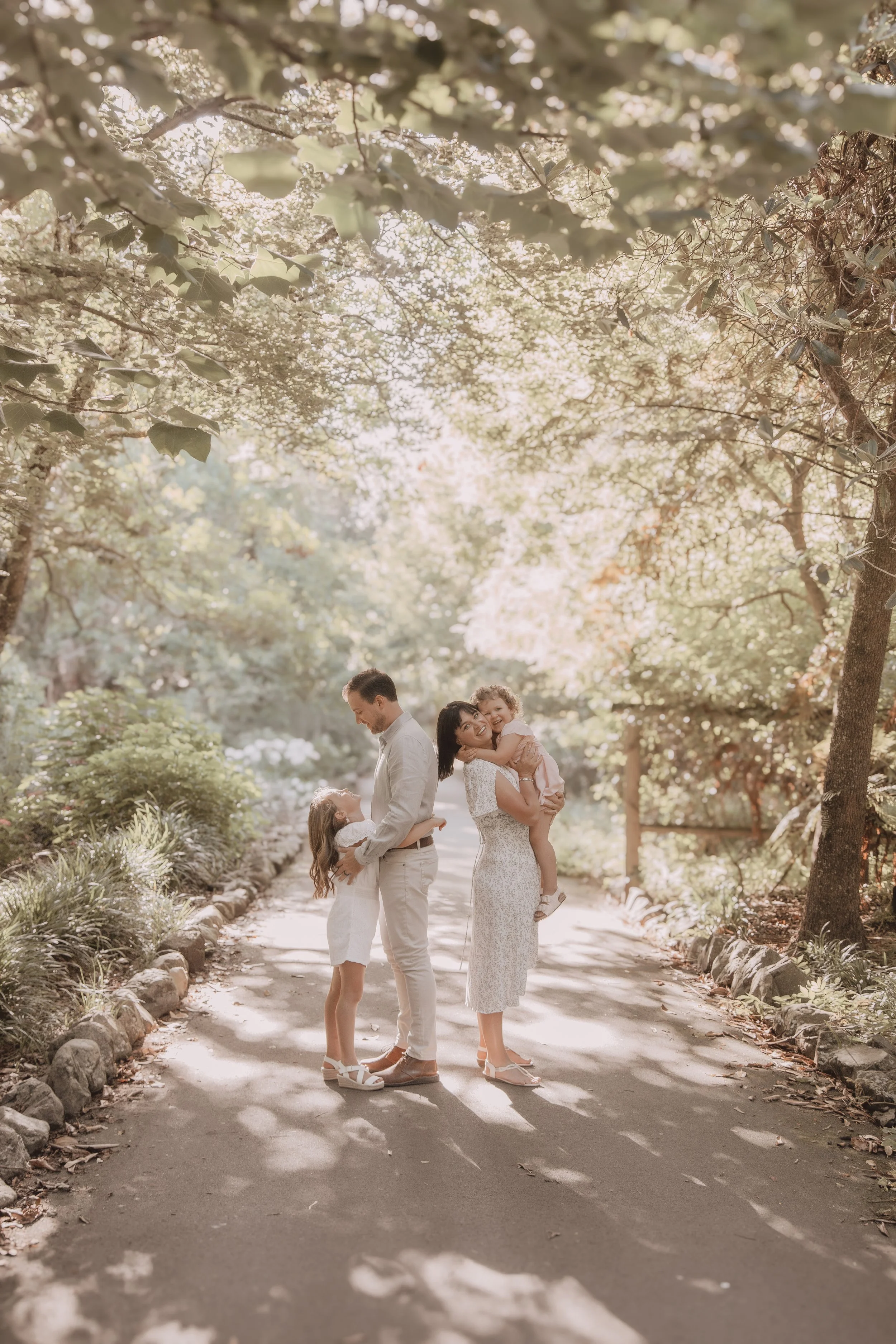 A family of four in a sunlit, tree-lined park, with two adults and two children, smiling and hugging, enjoying a warm day.
