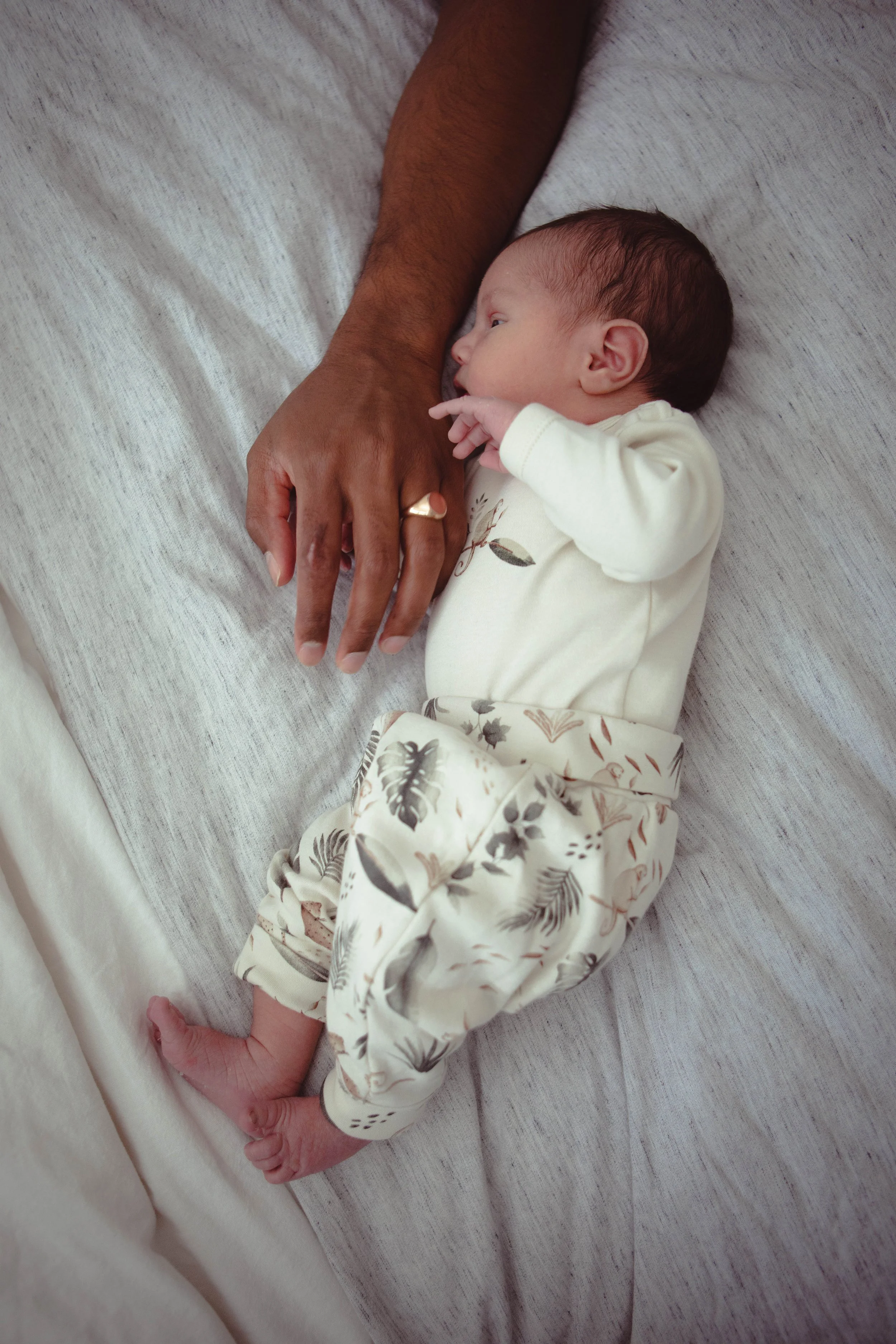 A baby laying on a bed, holding a person's arm with one hand, and looking at them. The person has a gold ring on their finger.