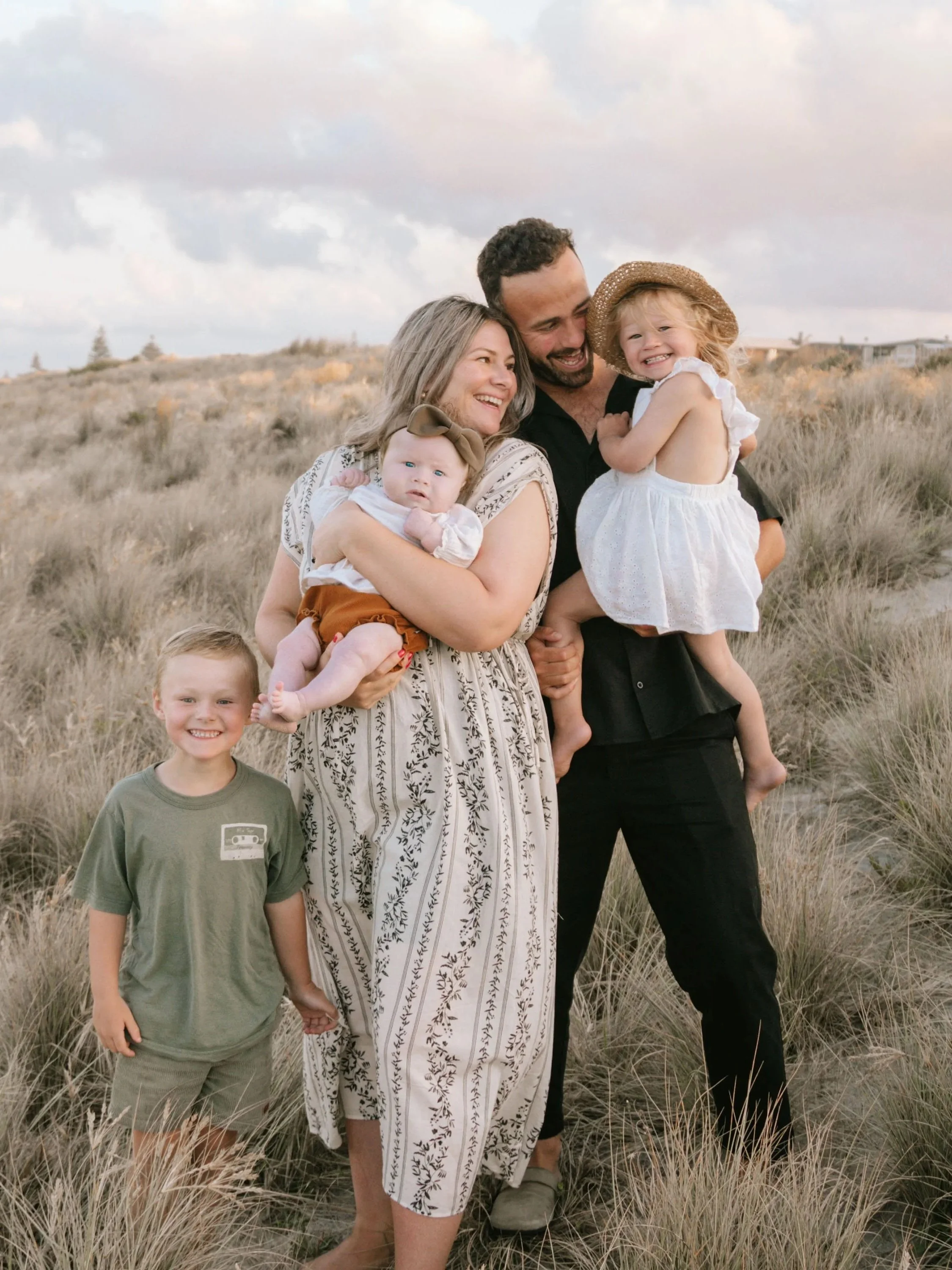 A family of five standing in a field of tall grass, smiling and enjoying each other's company during sunset. Two young children, a girl with a sun hat and a boy, along with their parents, a mother and father, are happily posing together.