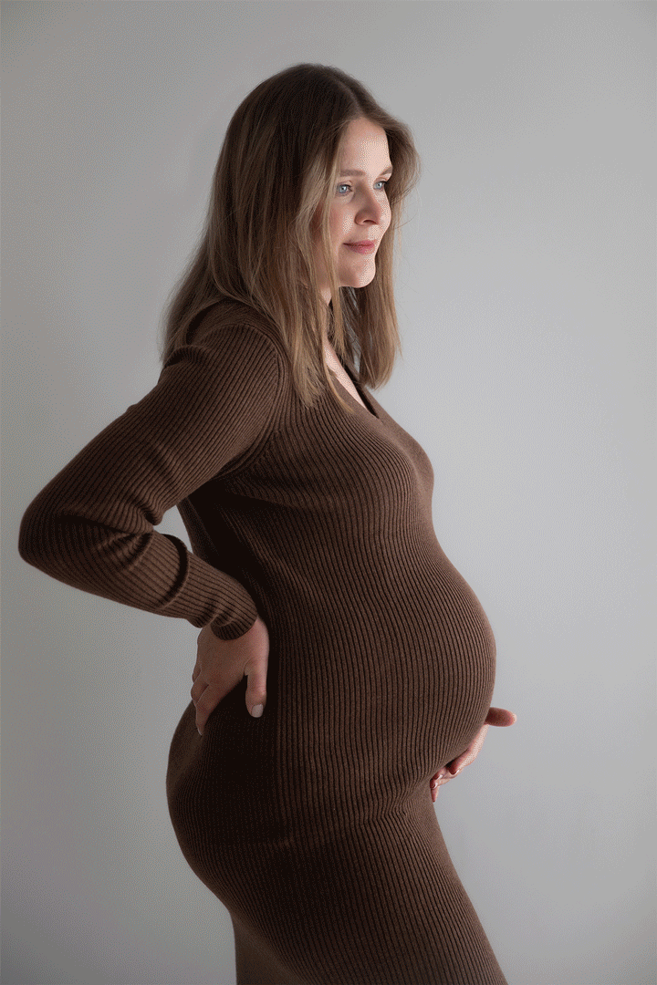 Pregnant woman in a fitted brown ribbed dress with long sleeves, standing sideways with one hand on her back and the other on her belly, looking thoughtfully to the side against a light grey background.
