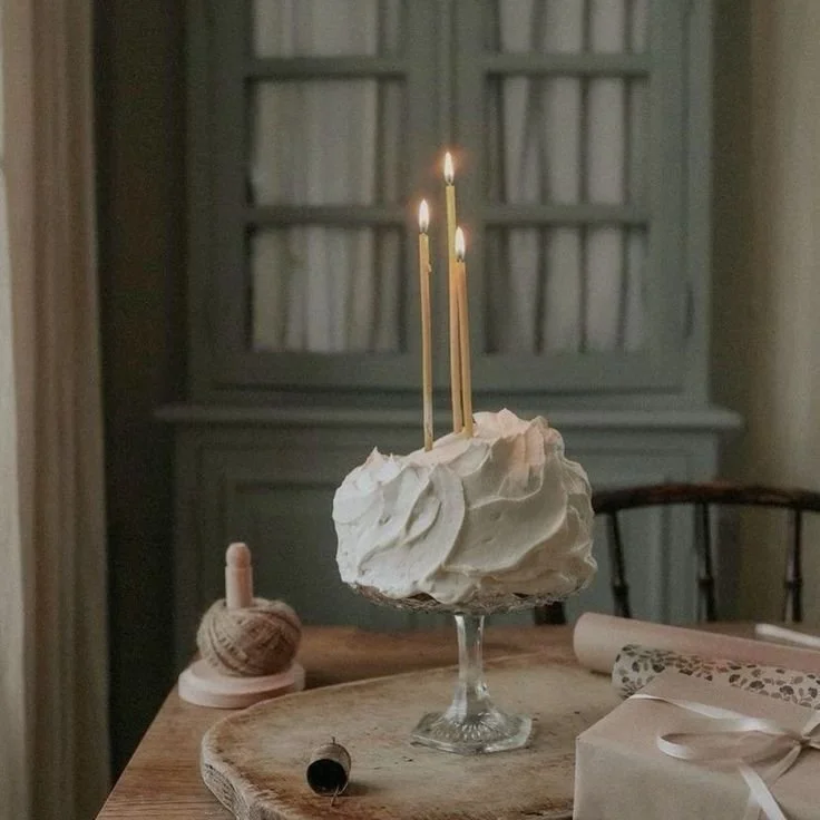A cake with white frosting on a glass stand with three lit candles on top, placed on a wooden table. Decorations include a small ball of yarn on a wooden base, a rolled paper or fabric, and a wrapped gift with a satin ribbon.
