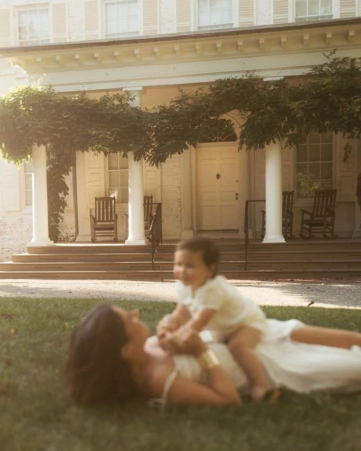 A woman and a child playing on the grass in front of a porch with white columns, rocking chairs, and a large door, with greenery overhead.