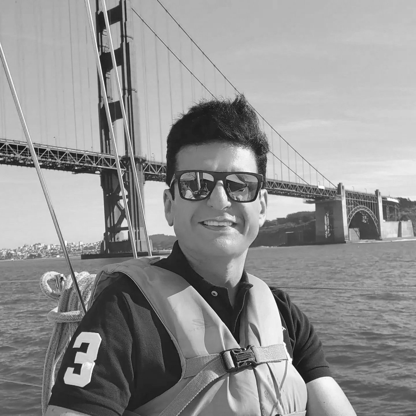 A man with dark hair and sunglasses smiling on a boat with the Golden Gate Bridge in the background.