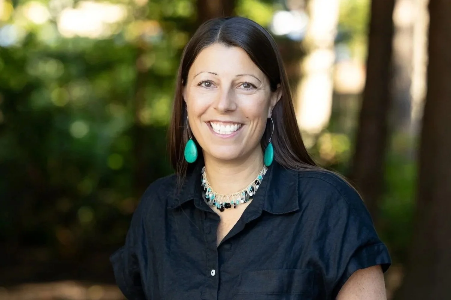 A woman with long brown hair smiling outdoors, wearing a black button-down short-sleeved top, a black and turquoise necklace, and dangling earrings, with a blurred green forest background.