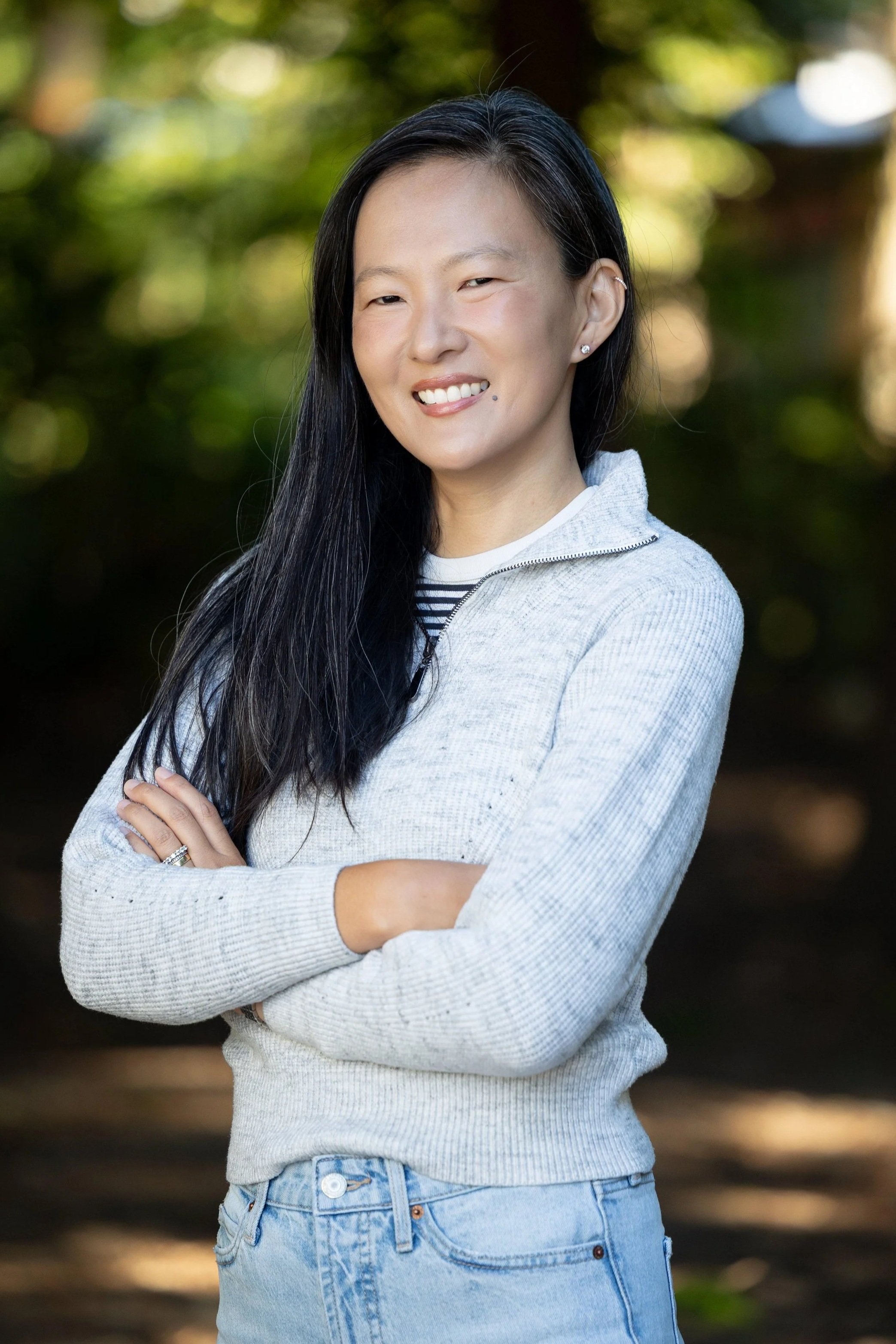 A woman with long, black hair wearing a gray sweater and posing with her arms crossed with a blurred forest background.