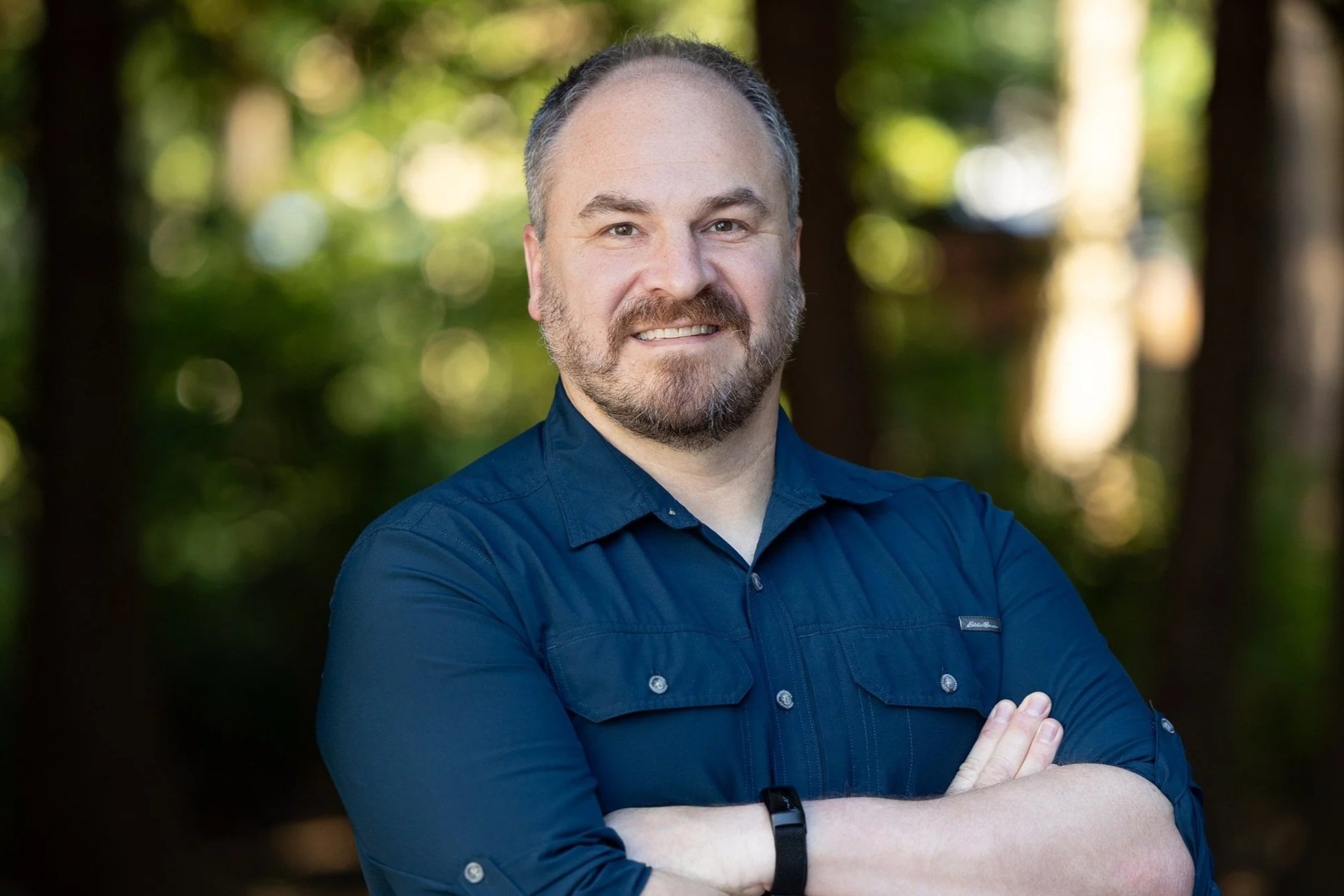 A man with a beard and short hair, wearing a light blue button-up shirt and watch, crosses his arms and smiles in an outdoor wooded area.