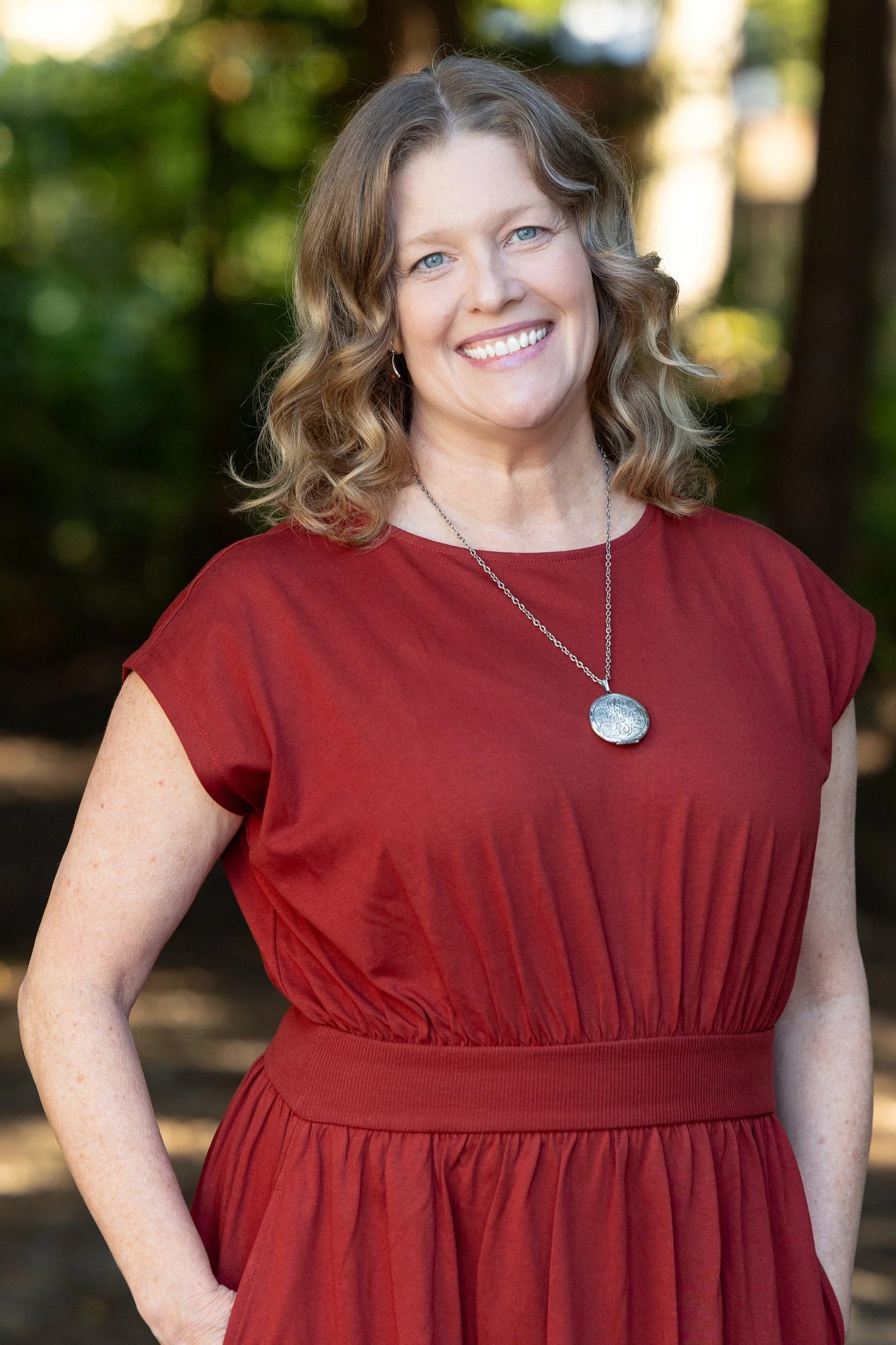 A smiling woman with red curly hair in a red dress standing in an outdoor setting with blurred trees in the background.