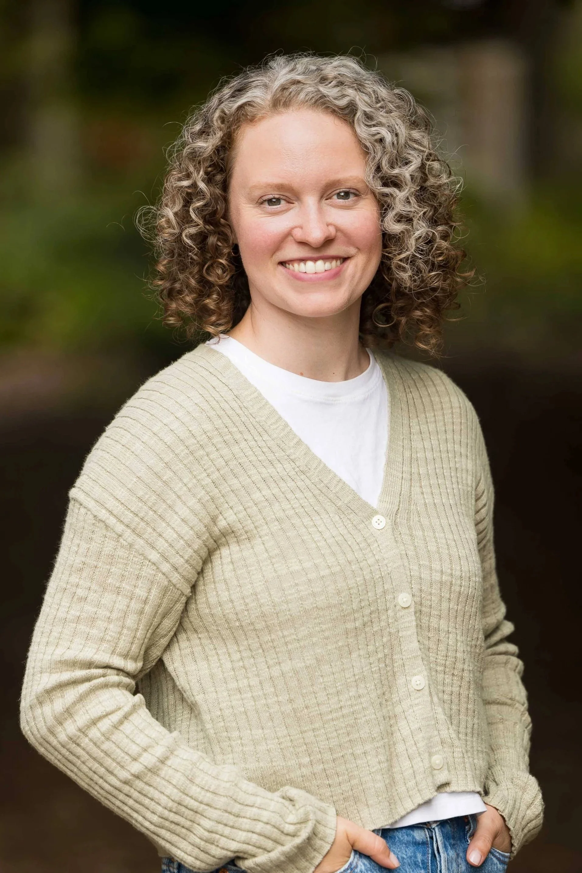 A woman with curly hair and a tan cardigan smiling with her hands in her pockets in front of a blurred forest background.