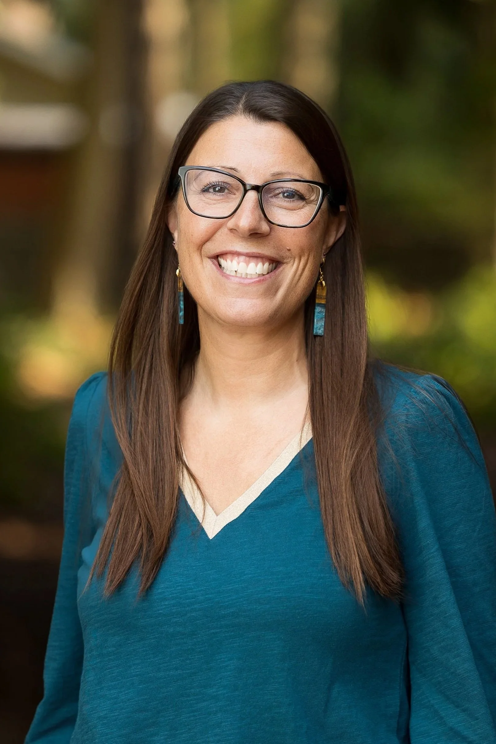 A woman with long brown hair smiling outdoors, wearing a white textured top, a black and turquoise necklace, and dangling earrings, with a blurred green forest background.