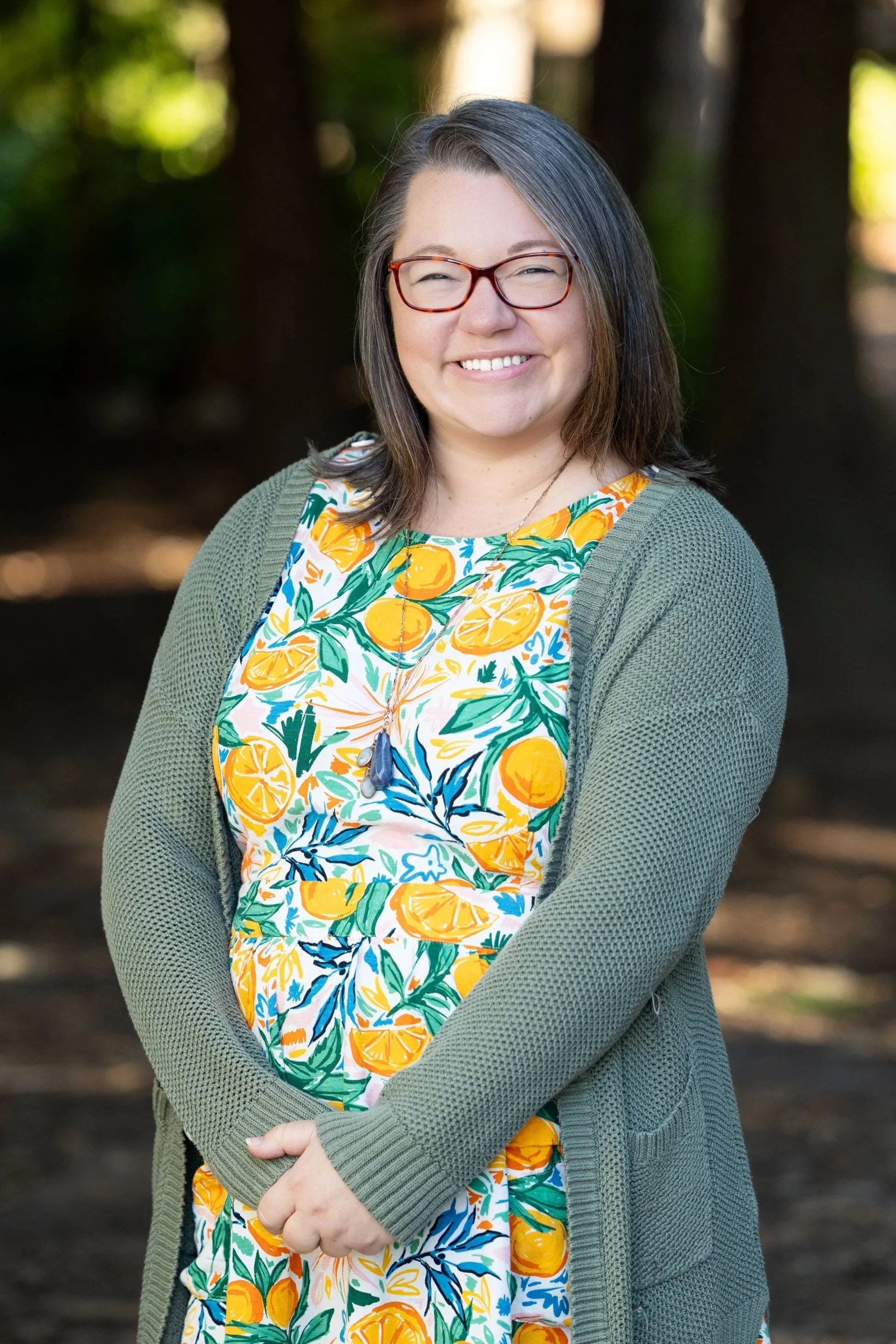 Woman with glasses and short dark hair smiling outdoors, wearing a colorful lemon print dress and a gray cardigan.