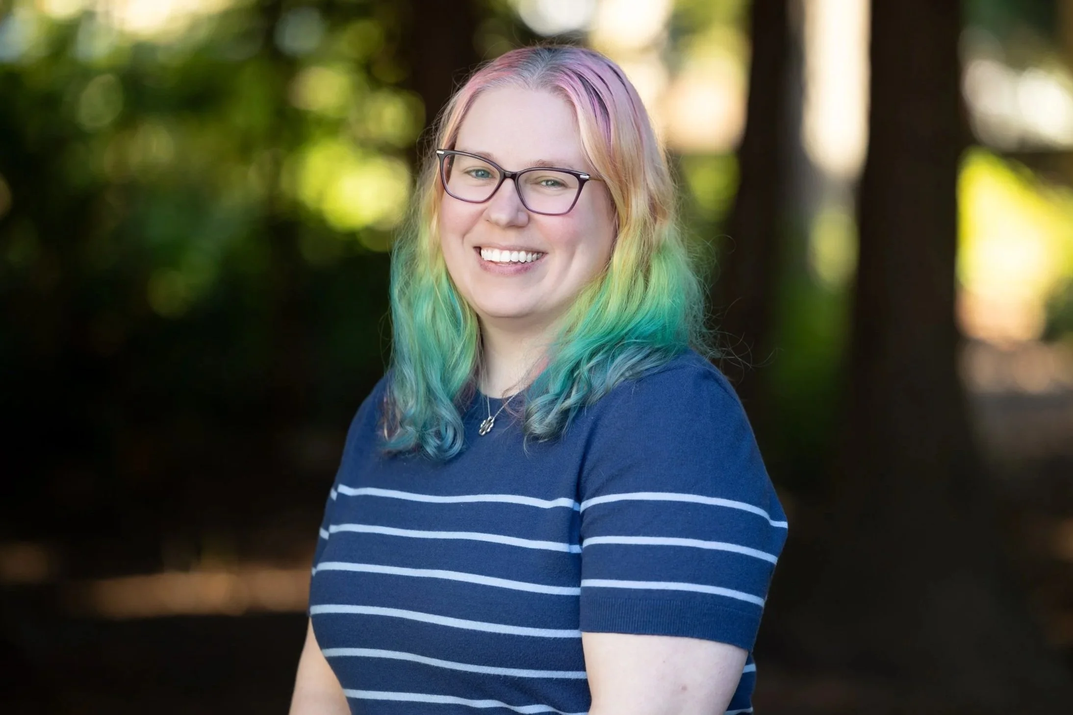 A young woman with rainbow hair, wearing glasses and a blue striped shirt, smiling outdoors in a forest setting.