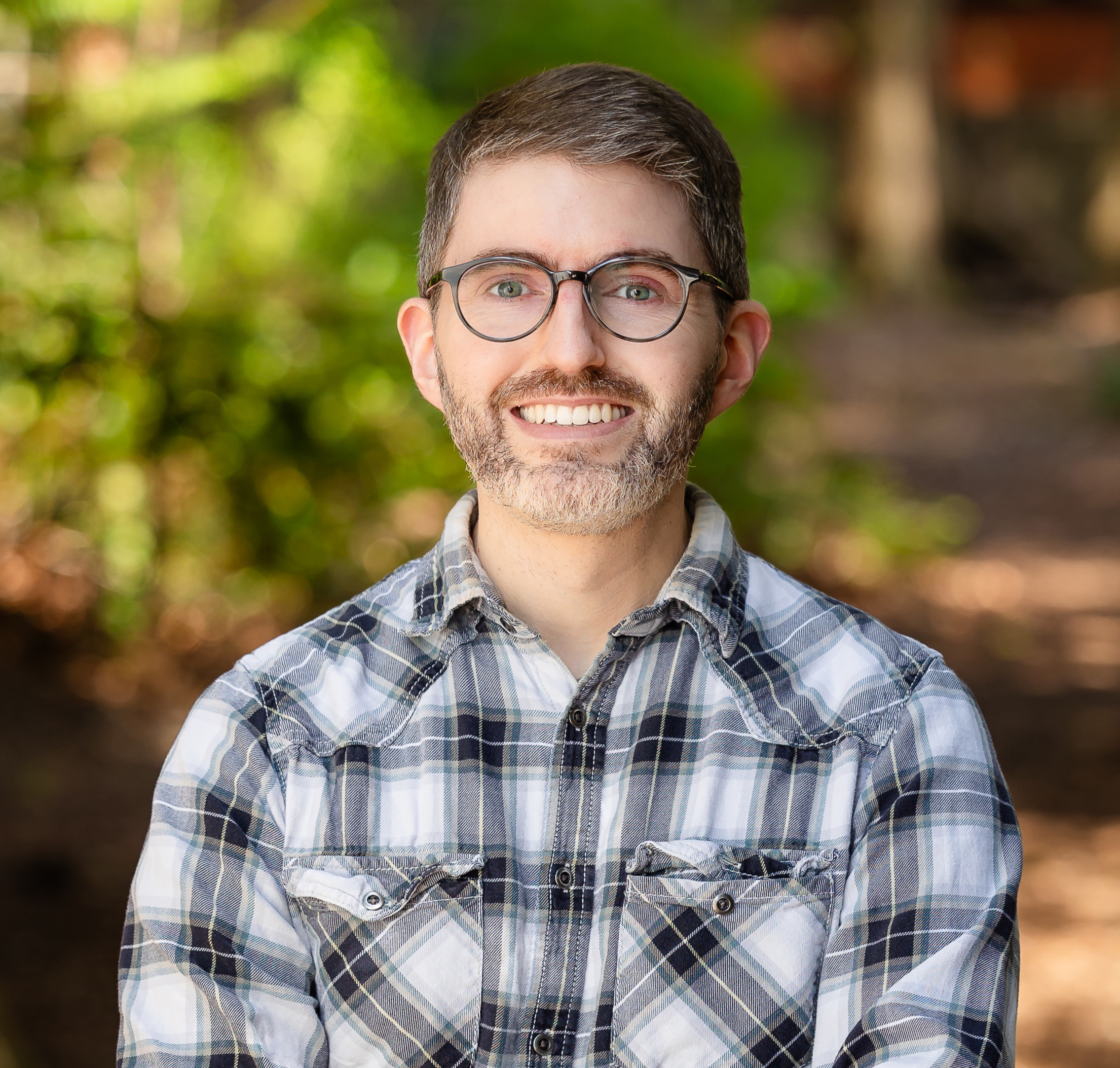 A man with short brown hair and glasses wearing a white and black plaid shirt standing in front of a wooded background that is blurred.