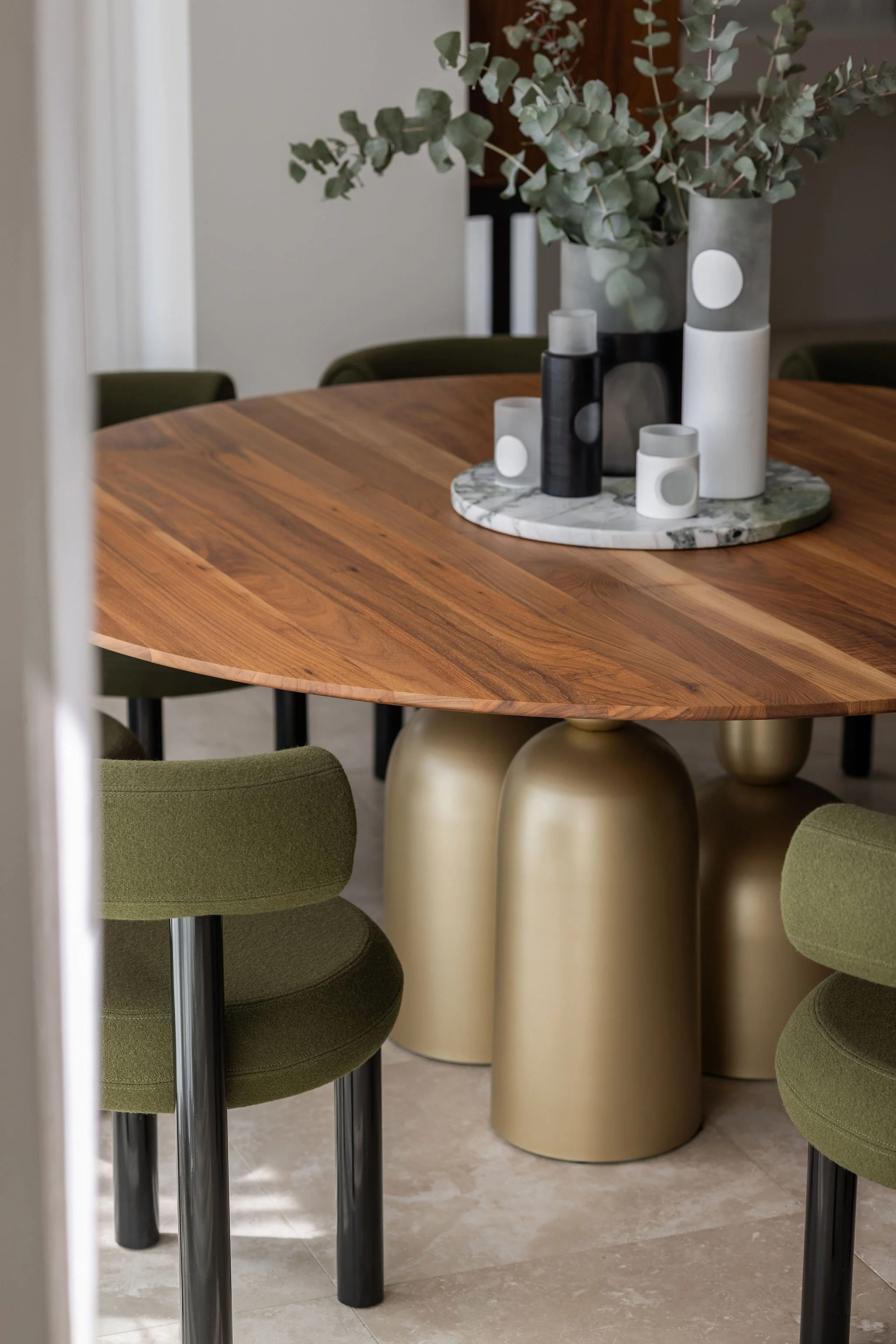 A round wooden table with a marble centerpiece decorated with tall vases and candles, surrounded by green upholstered chairs with metal legs, on a tiled floor.