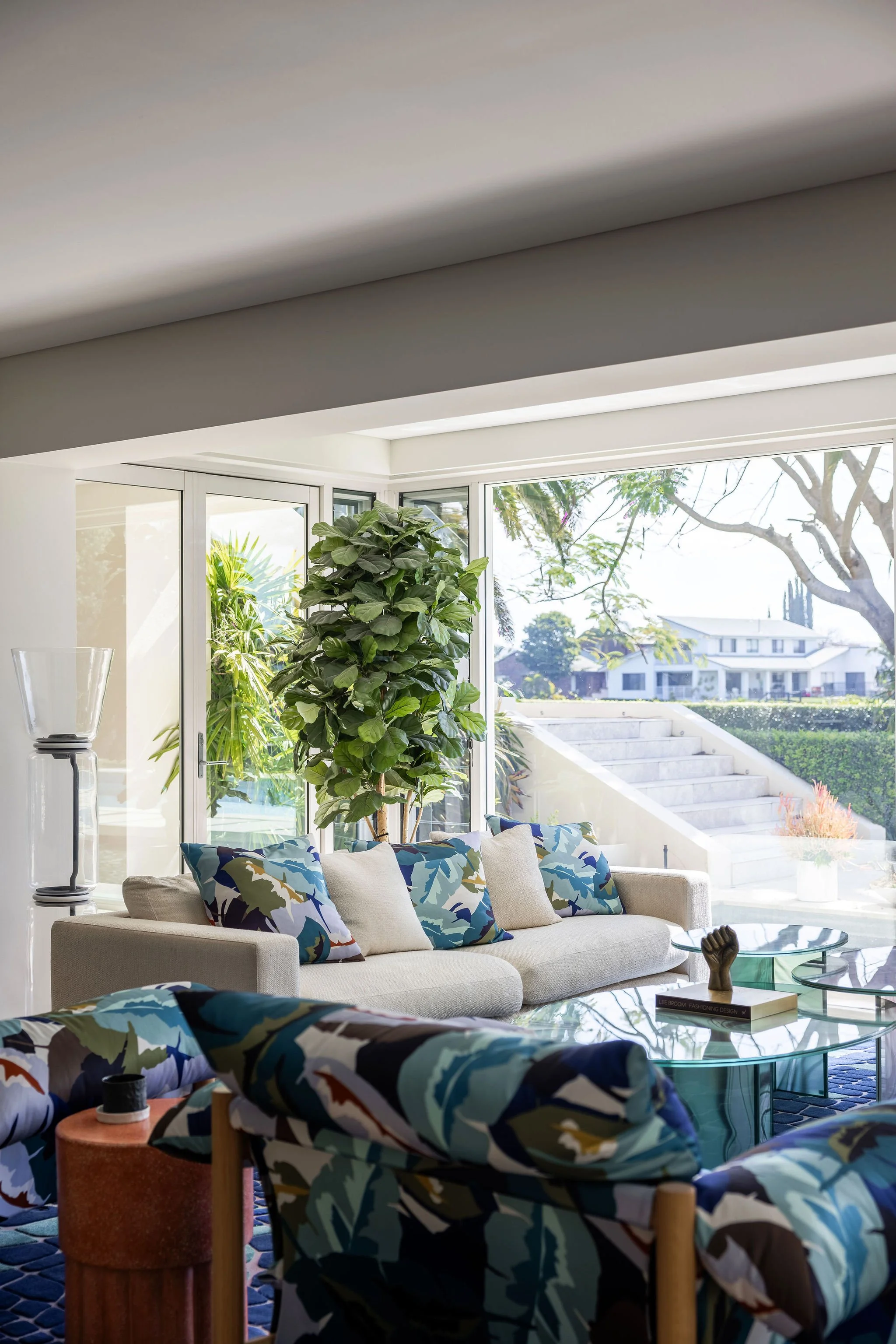 A bright living room with large glass windows showing outdoor trees and houses. There is a beige sofa with colorful abstract-patterned pillows, a glass coffee table, and a large potted plant near the window.