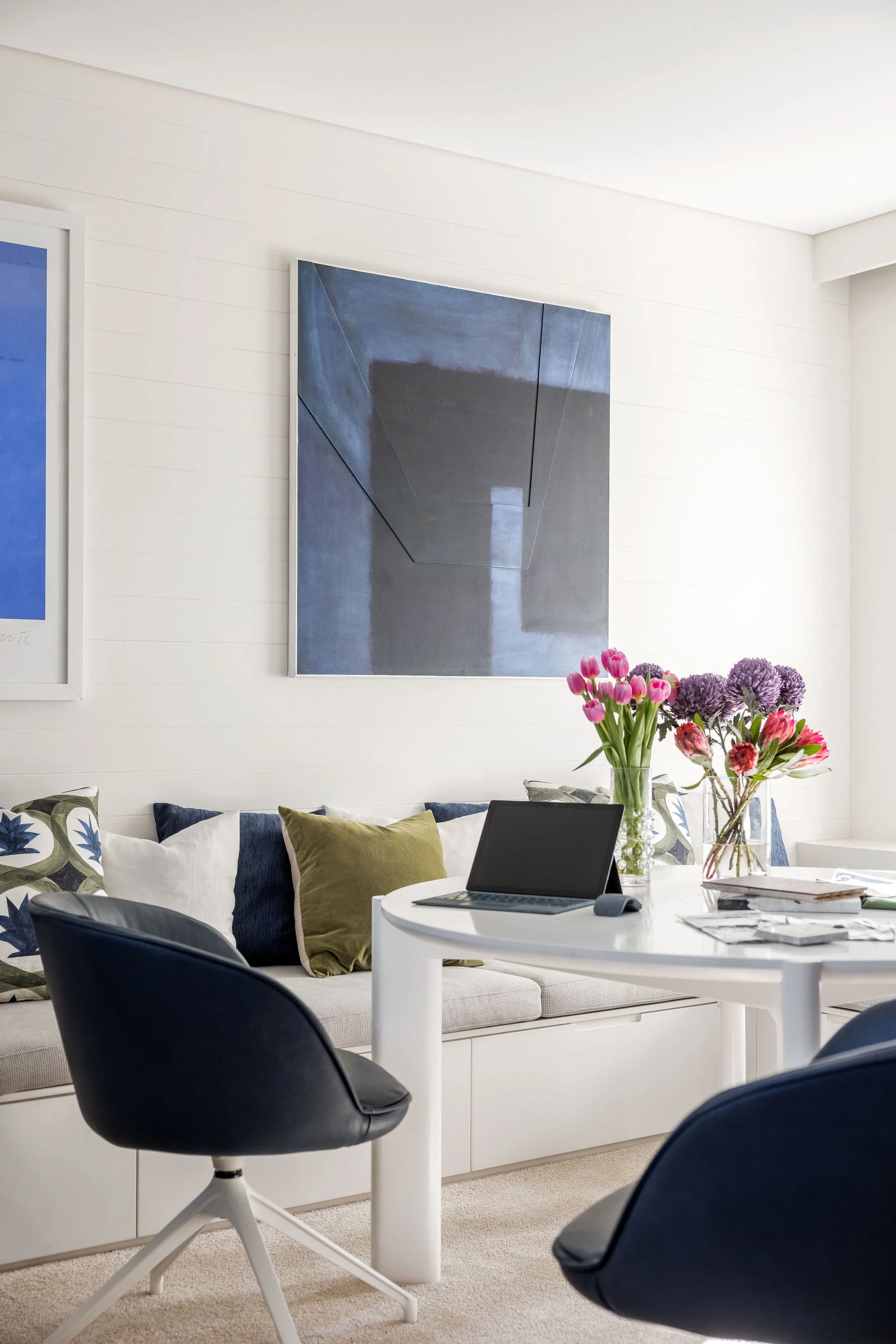 A modern Gold Coast living room with a white couch adorned with floral and solid-colored pillows, a round white table with an open laptop, a vase of pink tulips and purple flowers, a black chair, and wall art including a large abstract painting.