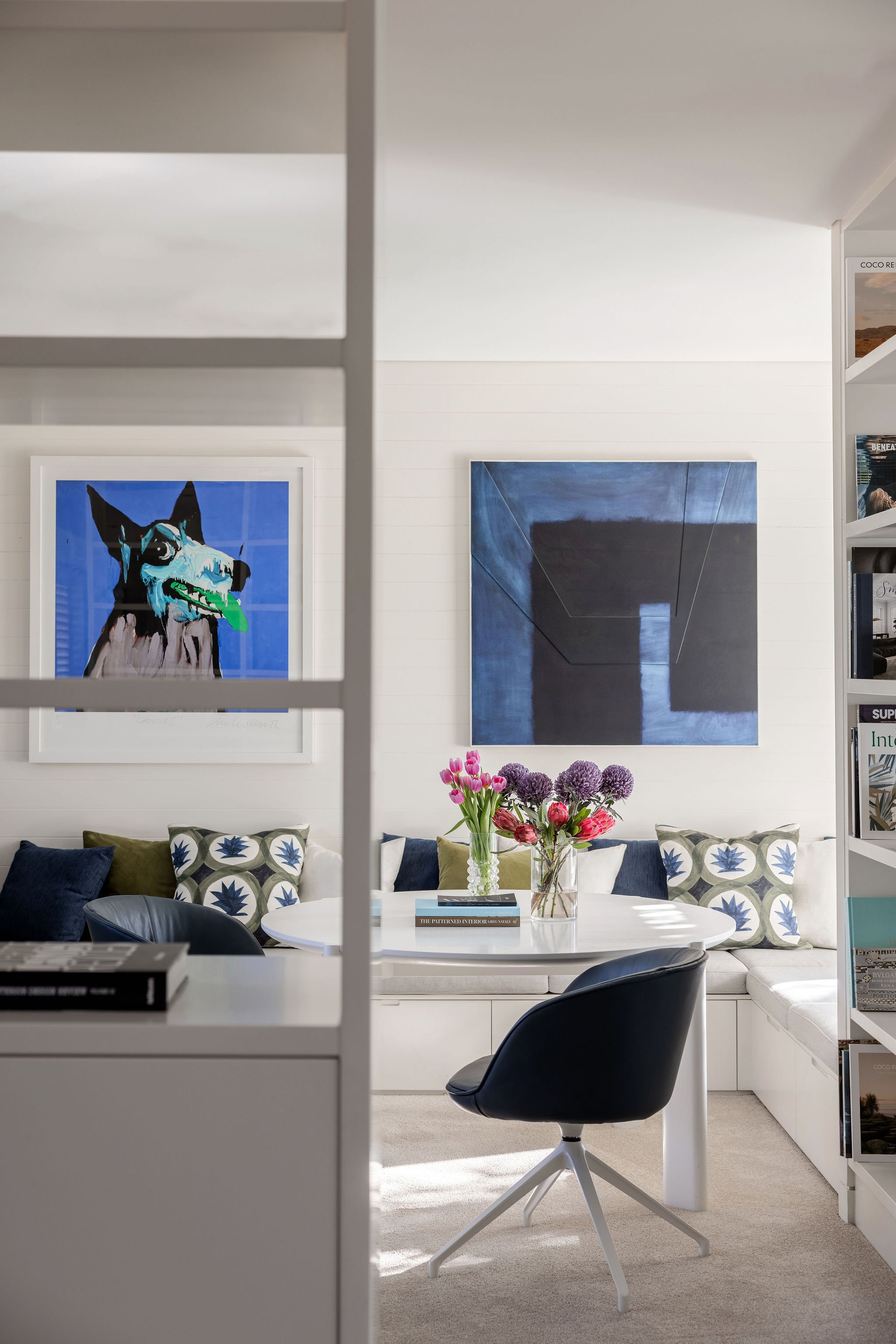 Modern Broadbeach Waters study with a white round table, black swivel chair, sofa with decorative pillows, vibrant floral arrangement, abstract artworks, books, and magazines.