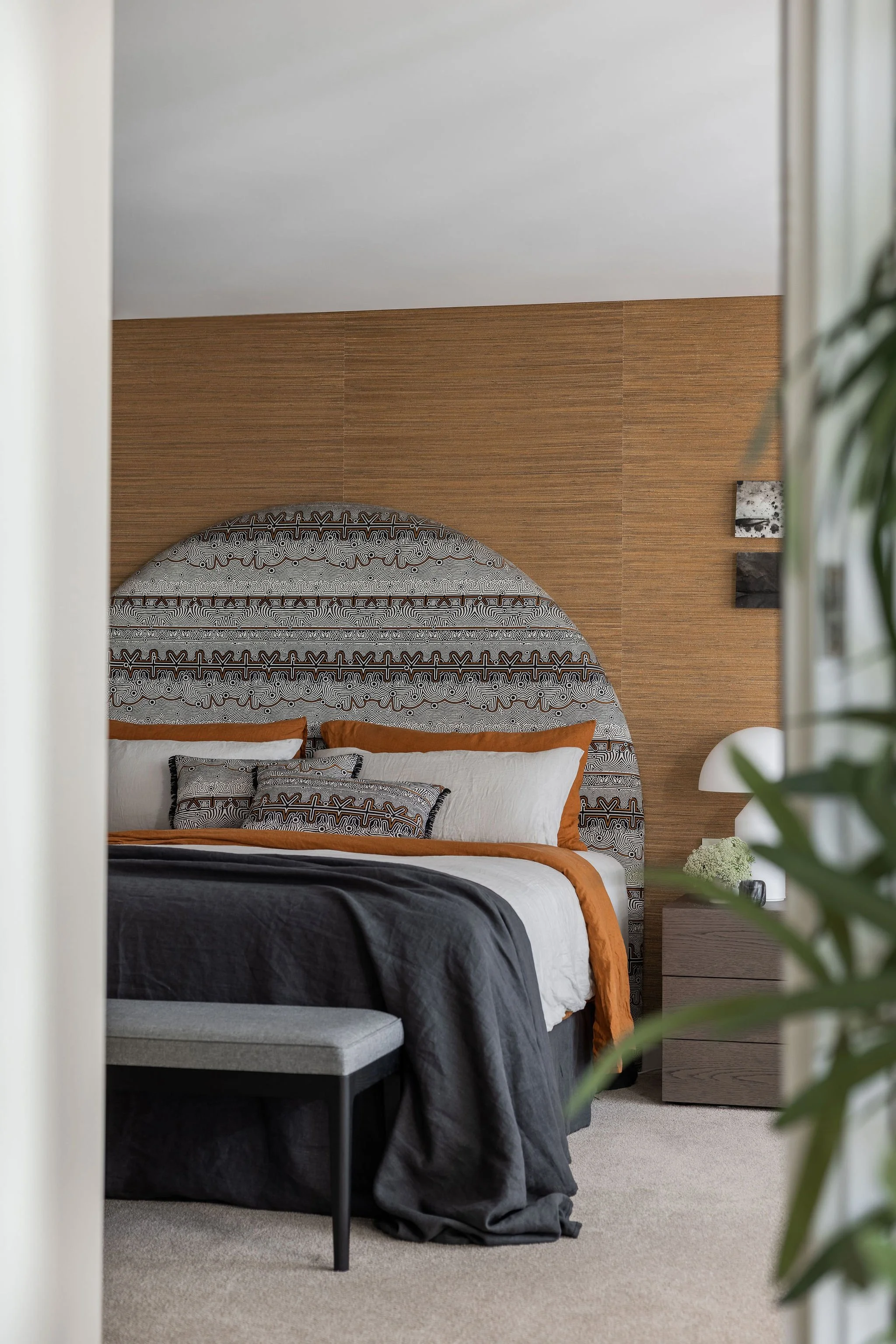 A bedroom with a bed featuring an upholstered headboard with black and white geometric patterns, brown accents, and white pillows. A dark gray blanket is draped over the bed. There is a wooden nightstand with a small white lamp and a decorative objec