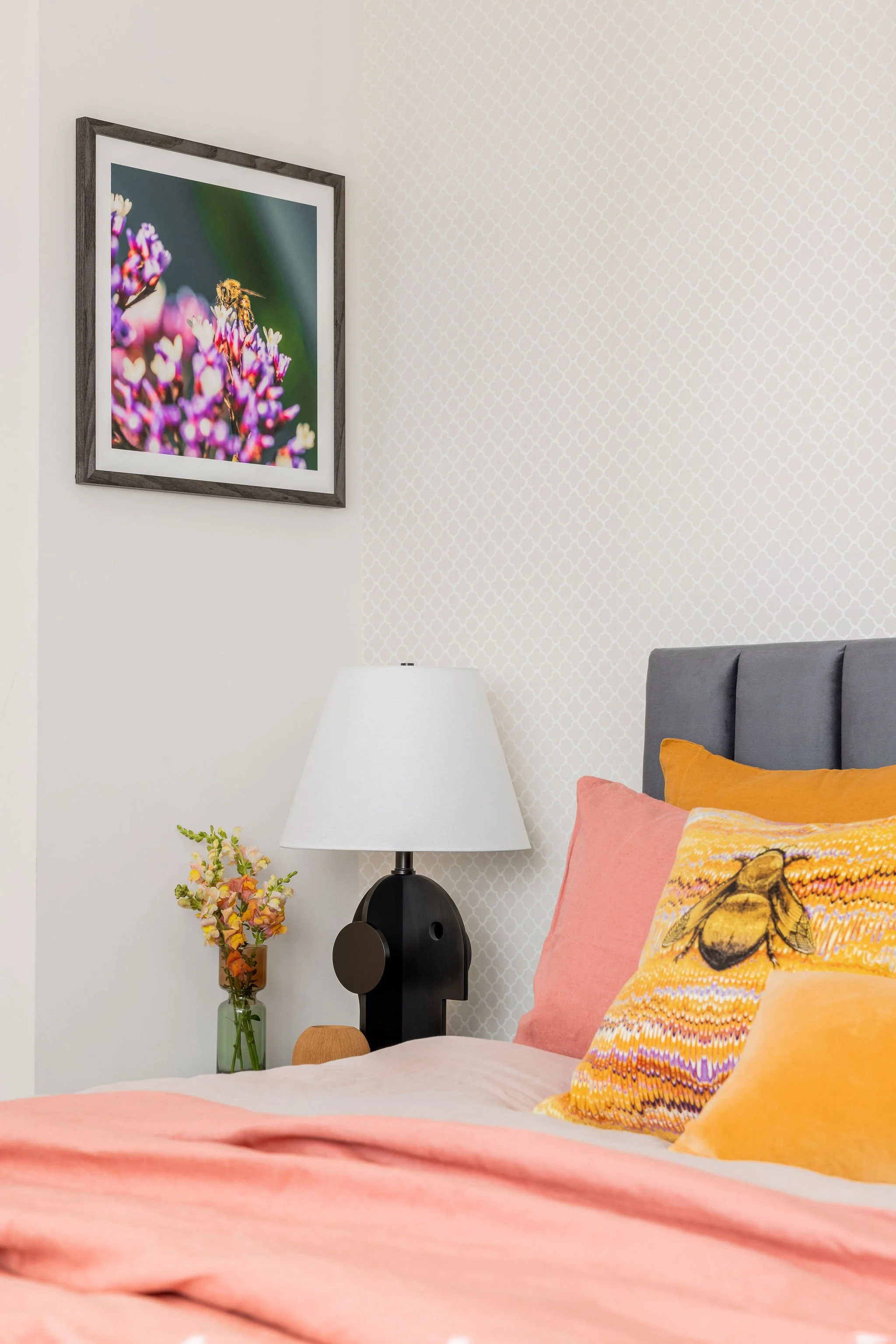A cozy Gold Coast bedroom corner with a bed dressed in pink, orange, and yellow pillows, a black bedside lamp, a tall green vase with flowers, a small wooden stool, and a framed close-up picture of a bee on purple flowers on the wall.