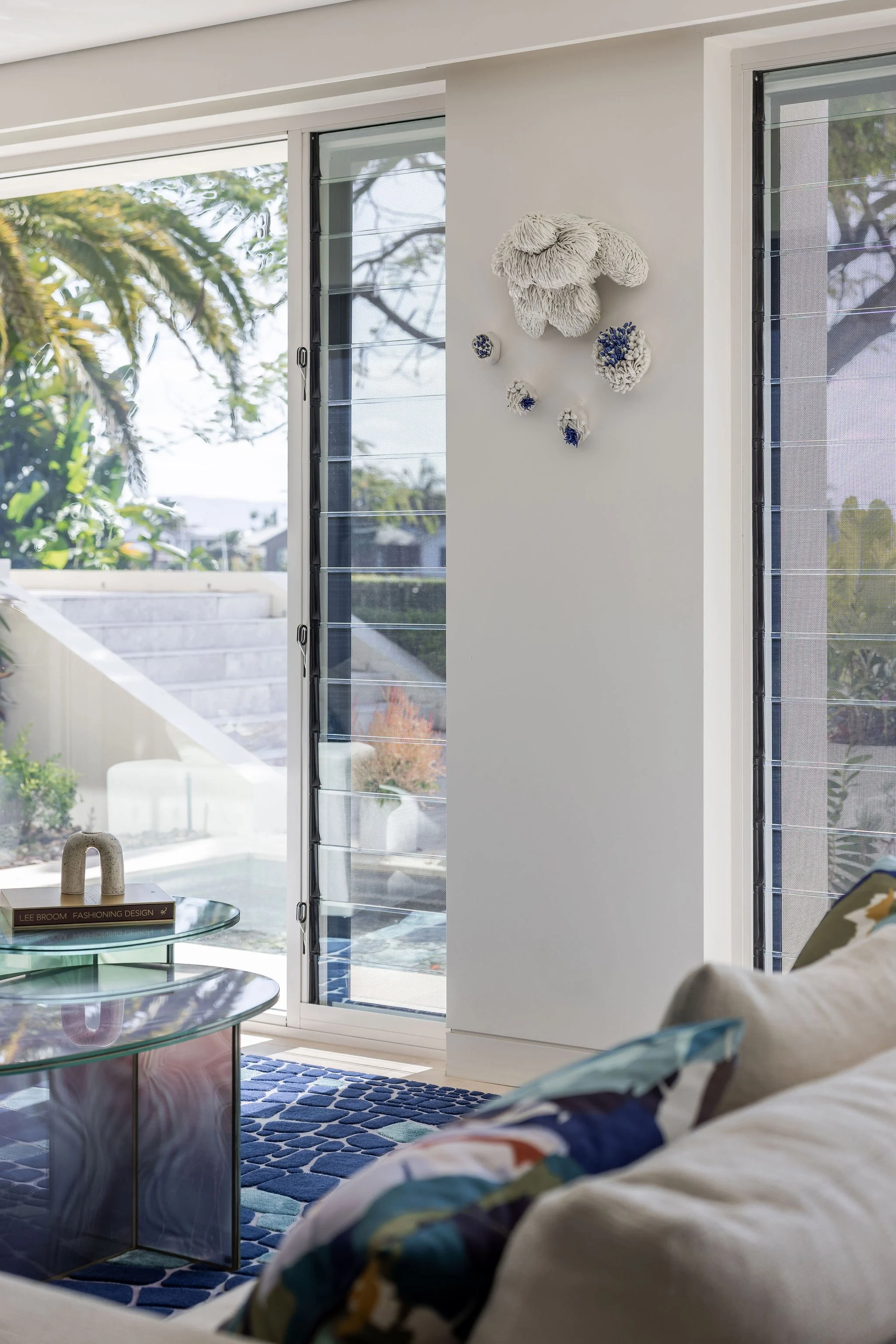 Interior of a modern living room with large glass sliding doors, a blue patterned rug, a glass coffee table, and a white wall with textured wall art. Outdoors, green trees and a staircase are visible.