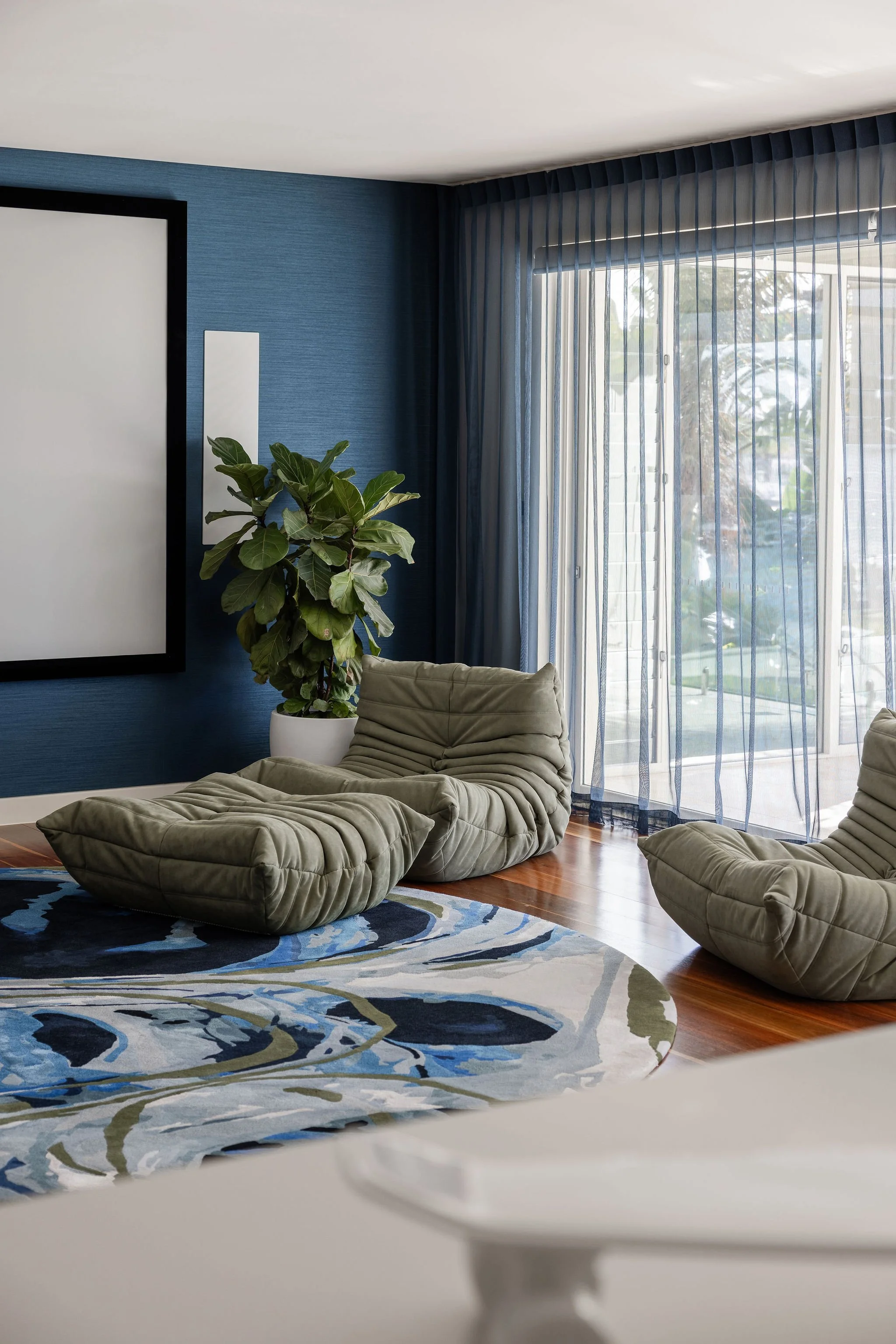 Contemporary Gold Coast cinema room with blue textured wall, large window with sheer curtains, green potted plant, patterned area rug, and modern green lounge chairs.