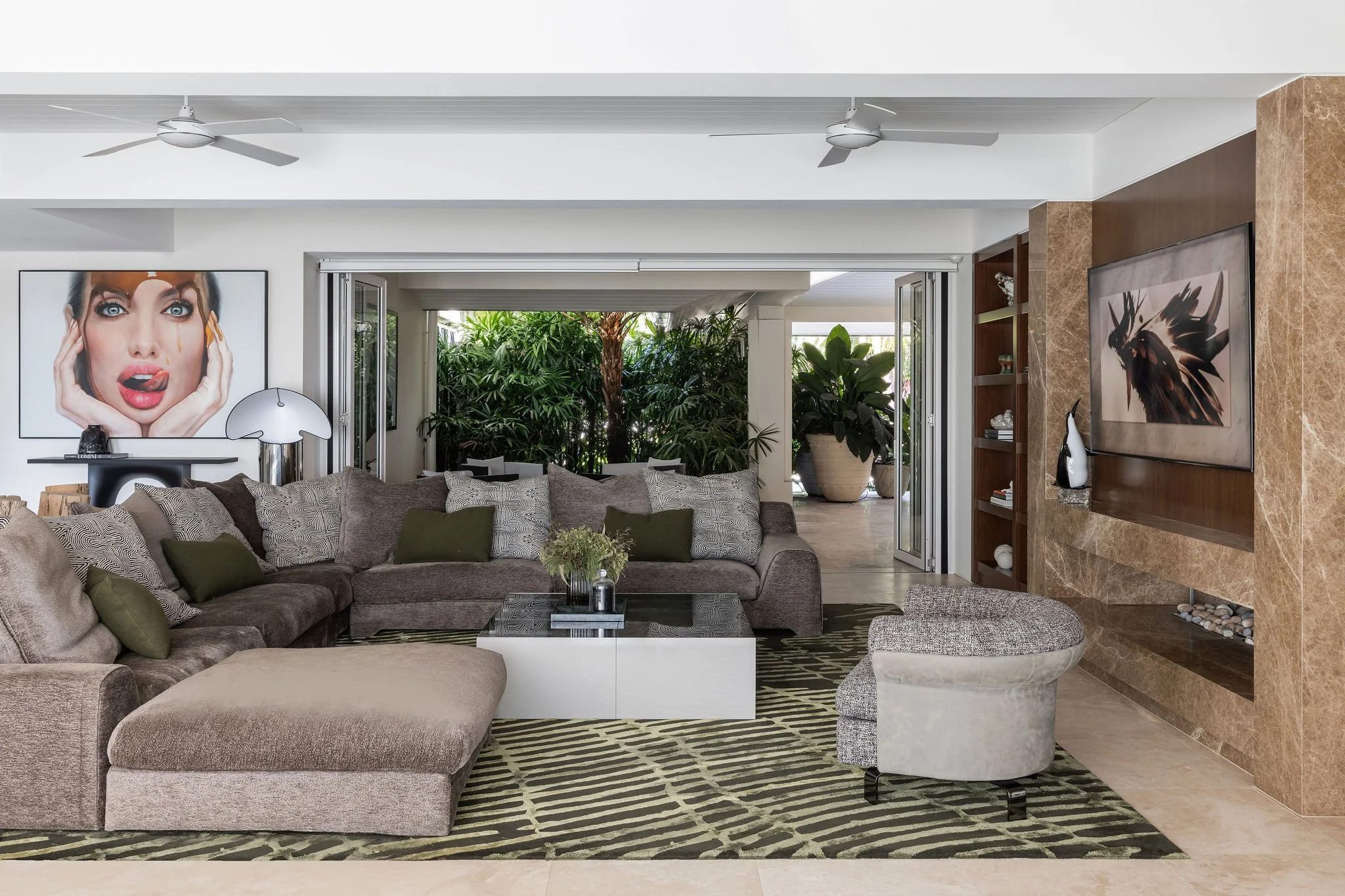 Interior of a modern Gold Coast living room with a large L-shaped Jardan Vista couch, decorative Kelly Werstler pillows, a square coffee table with a small plant, ceiling fans, and large sliding glass doors opening to a lush garden.