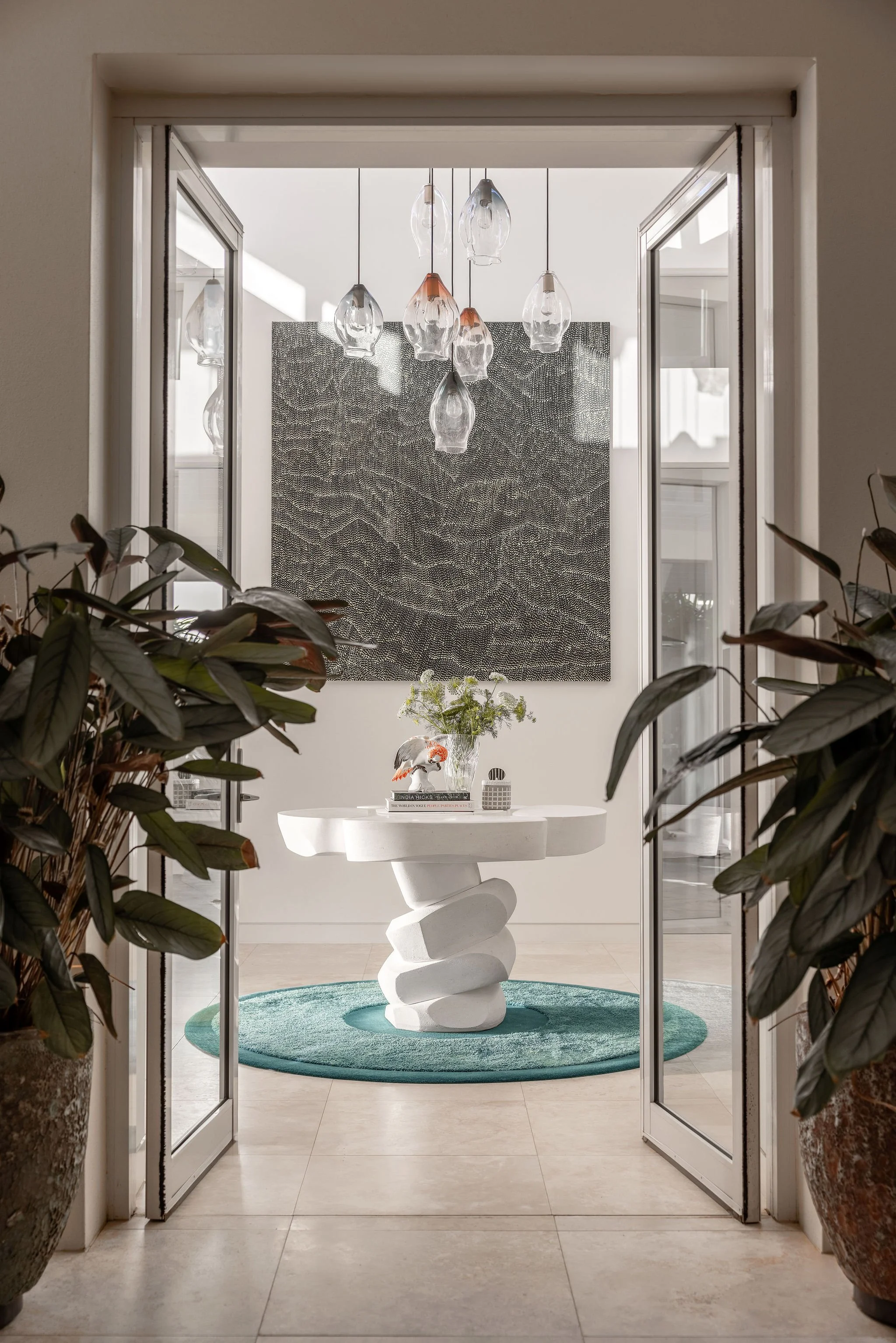 Living room with large rectangular wall art, hanging glass pendant lights, white sculptural table on a blue circular rug, surrounded by large potted plants.