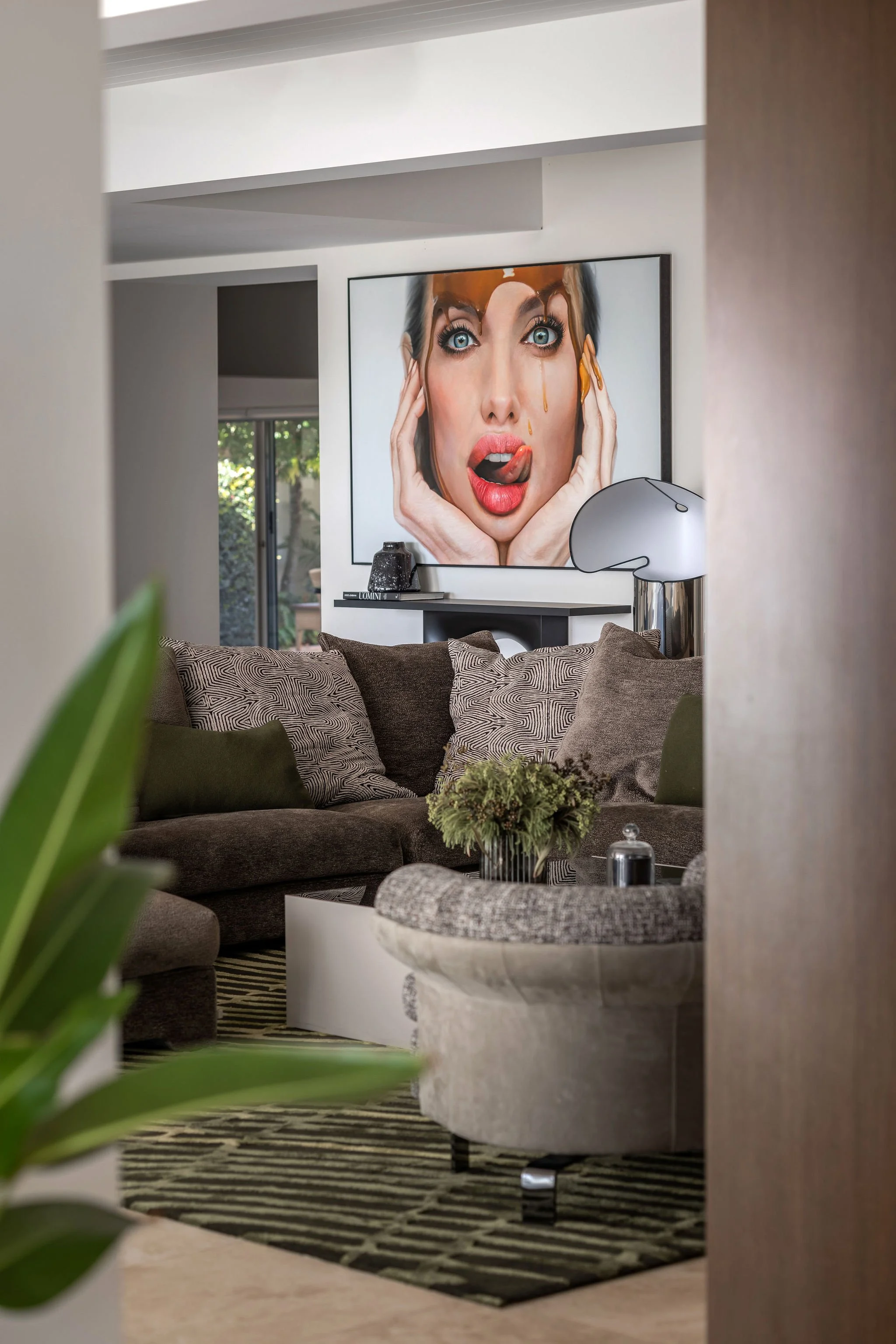 Living room with a large framed portrait of a woman with bright blue eyes and red lips hanging on the wall. The room contains a brown sofa with patterned pillows, a dark table with decorative objects, and a black-and-white striped rug.
